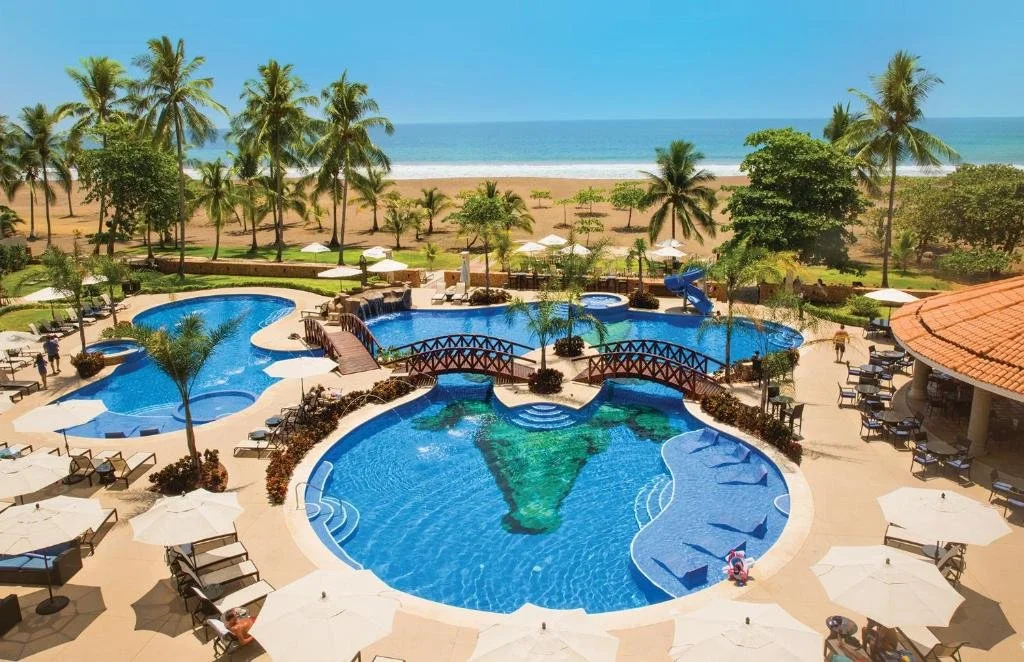 Resort pool area with multiple pools, surrounded by lounge chairs and umbrellas, with palm trees and a beach in the background.