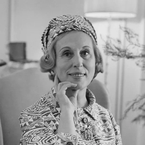 Black and white photo of a woman with a patterned headscarf and matching clothing, sitting indoors with a lamp and houseplant in the background.
