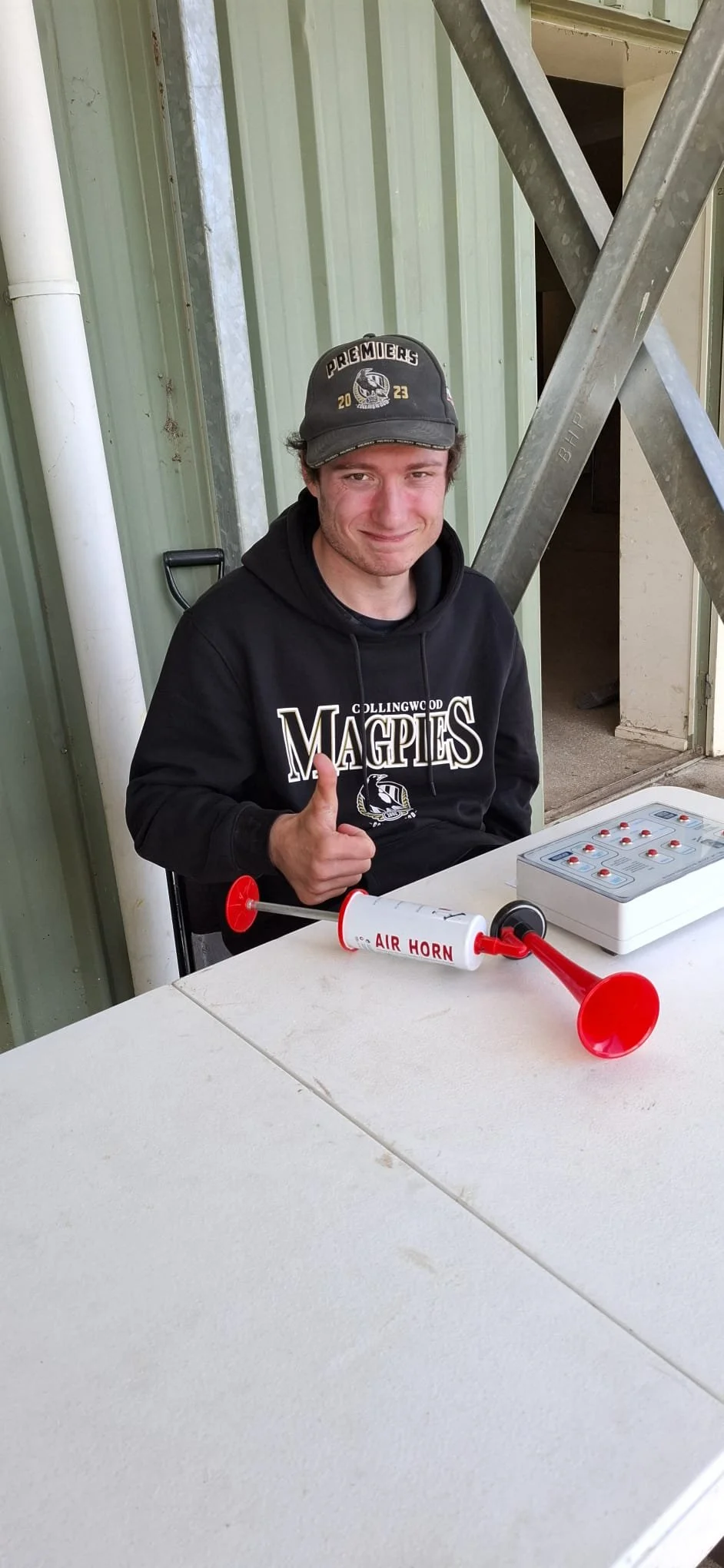A young man sitting at a table, giving a thumbs-up, with an air horn, a control panel, and a D-shaped object on the table.