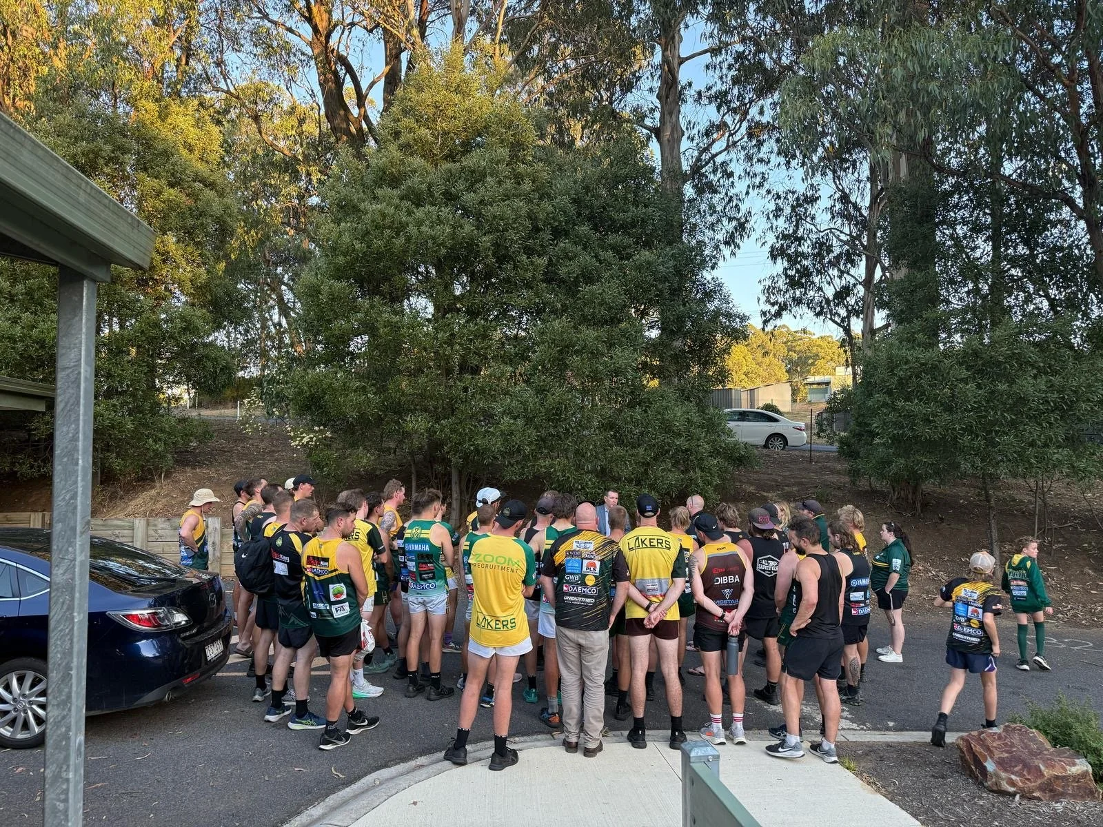 Group of athletes, mostly men, gathered outdoors for a team meeting or briefing, dressed in sports jerseys and shorts, with some wearing hats and sunglasses, surrounded by trees and parked cars.