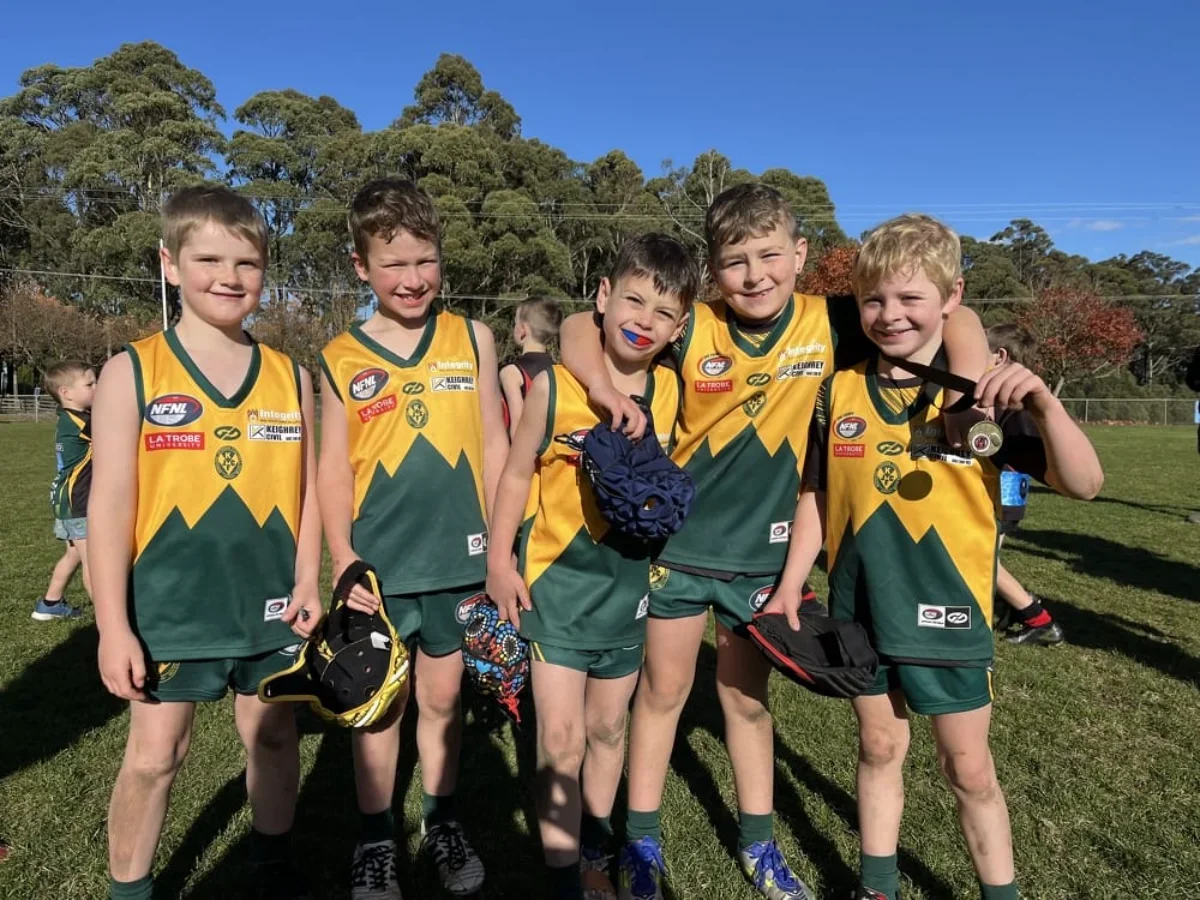 Five young boys in yellow and green sports uniforms, standing on a grassy field with trees in the background. One boy is holding a medal, and another has face paint. They are smiling and appear to be celebrating a sports event.