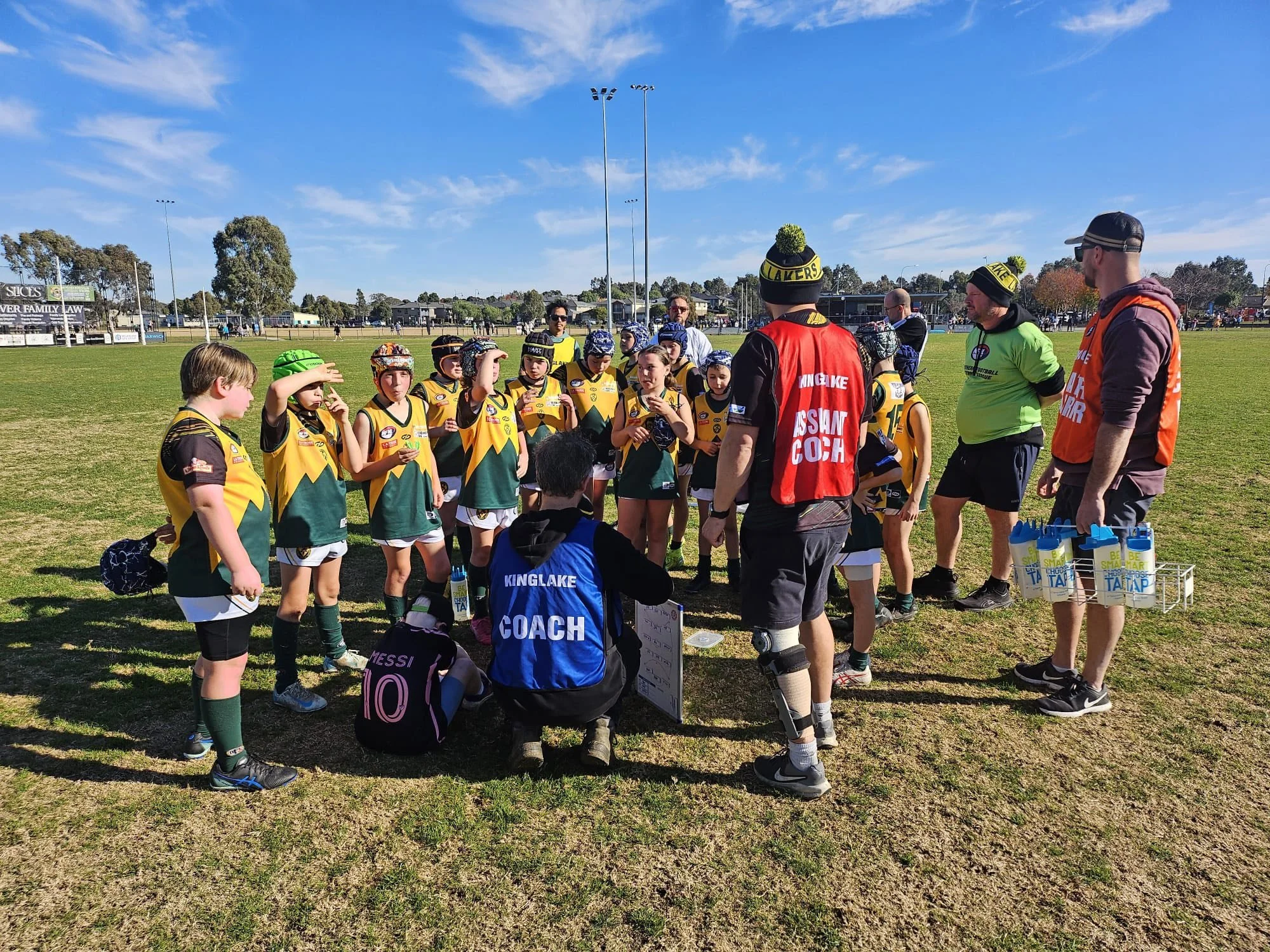 Kids sports team gathered around coaches on a grassy field during daytime practice or game, with bright sunny weather.
