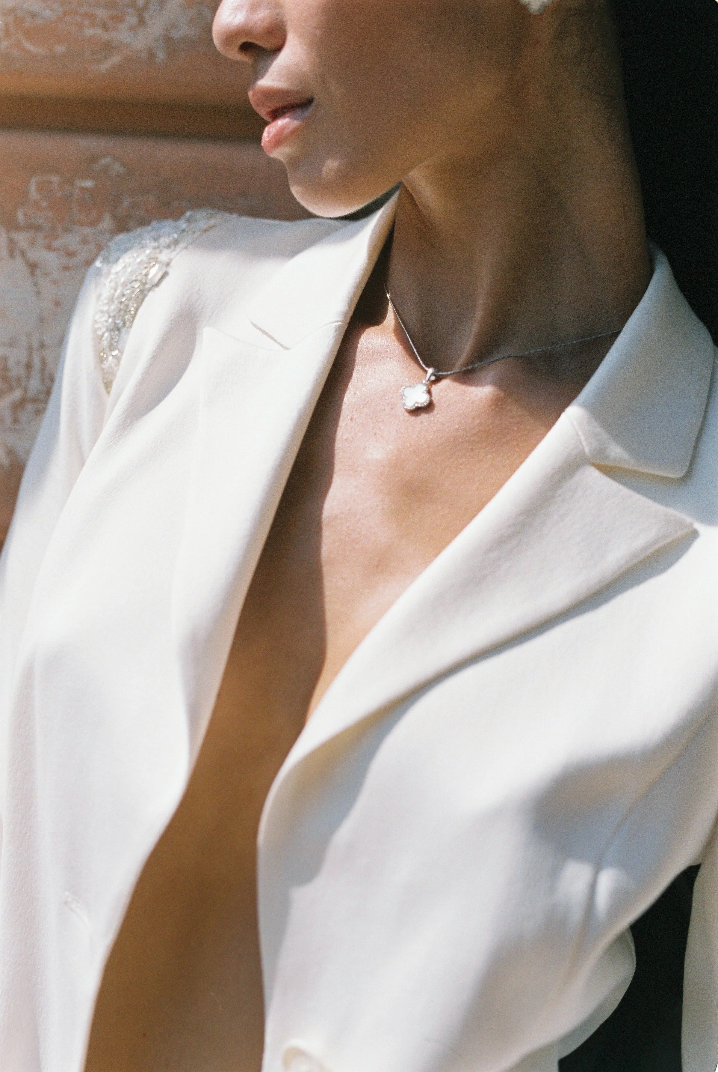 Close-up of a woman wearing a white blazer with embroidered shoulder details, a delicate necklace with a four-leaf clover pendant, and with her lips slightly parted, facing left.