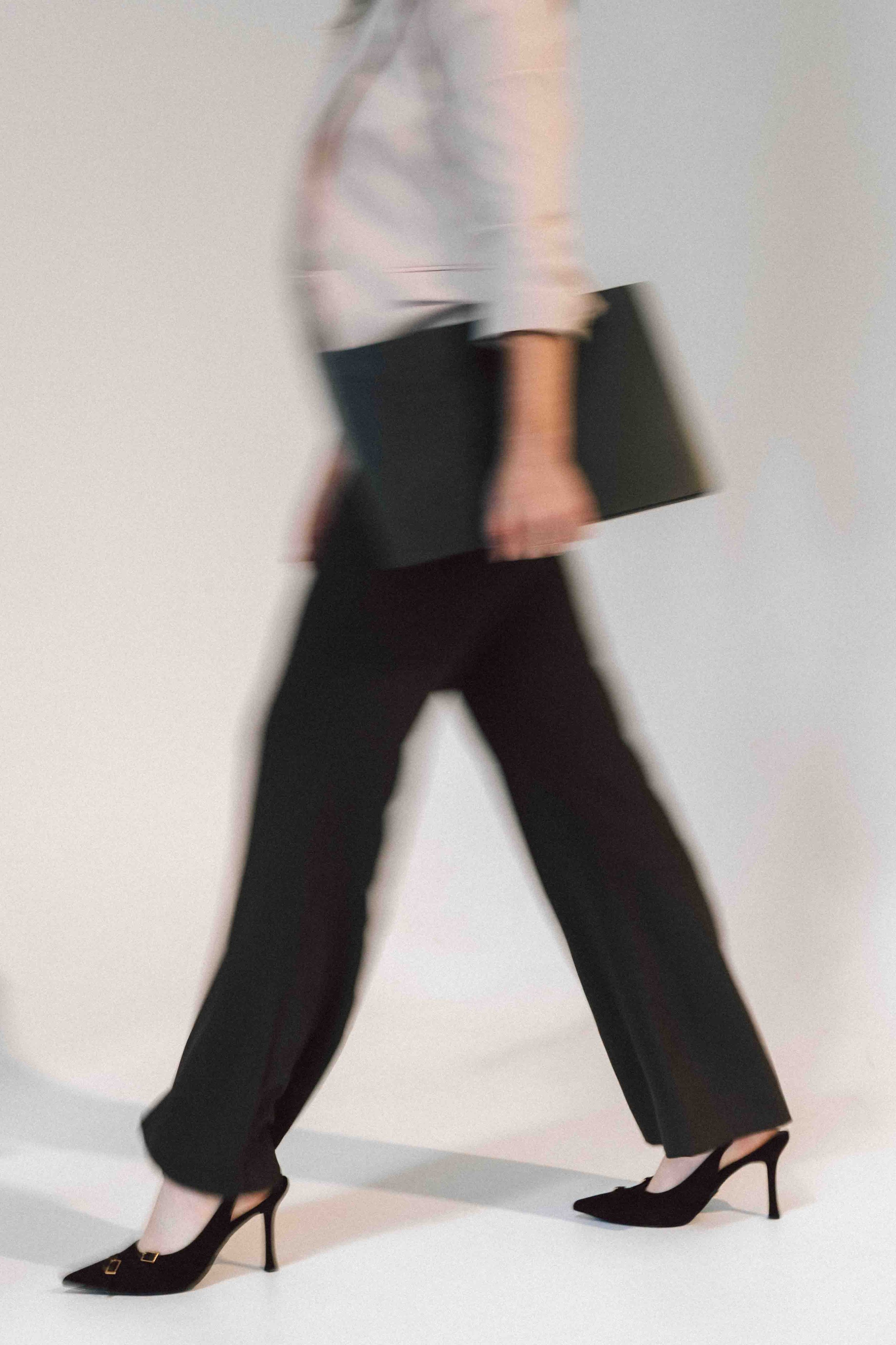 A woman walking with a blurred motion effect, dressed in black pants, a light-colored blazer, wearing black high heels, and carrying a black folder or clutch.