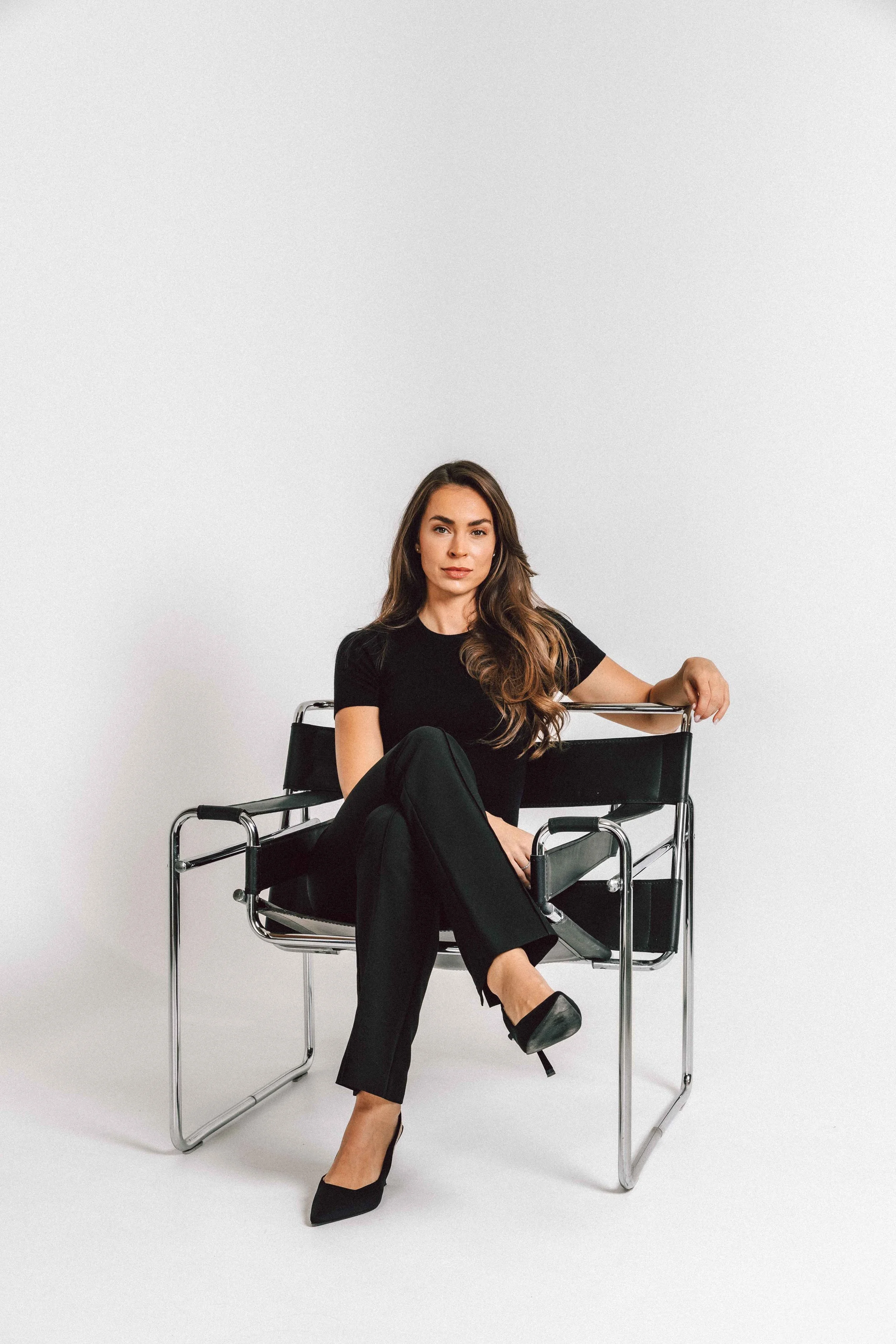 A woman sitting on a modern black chair with chrome frame against a plain white background. She has long brown hair, wearing a black outfit and black high heels.