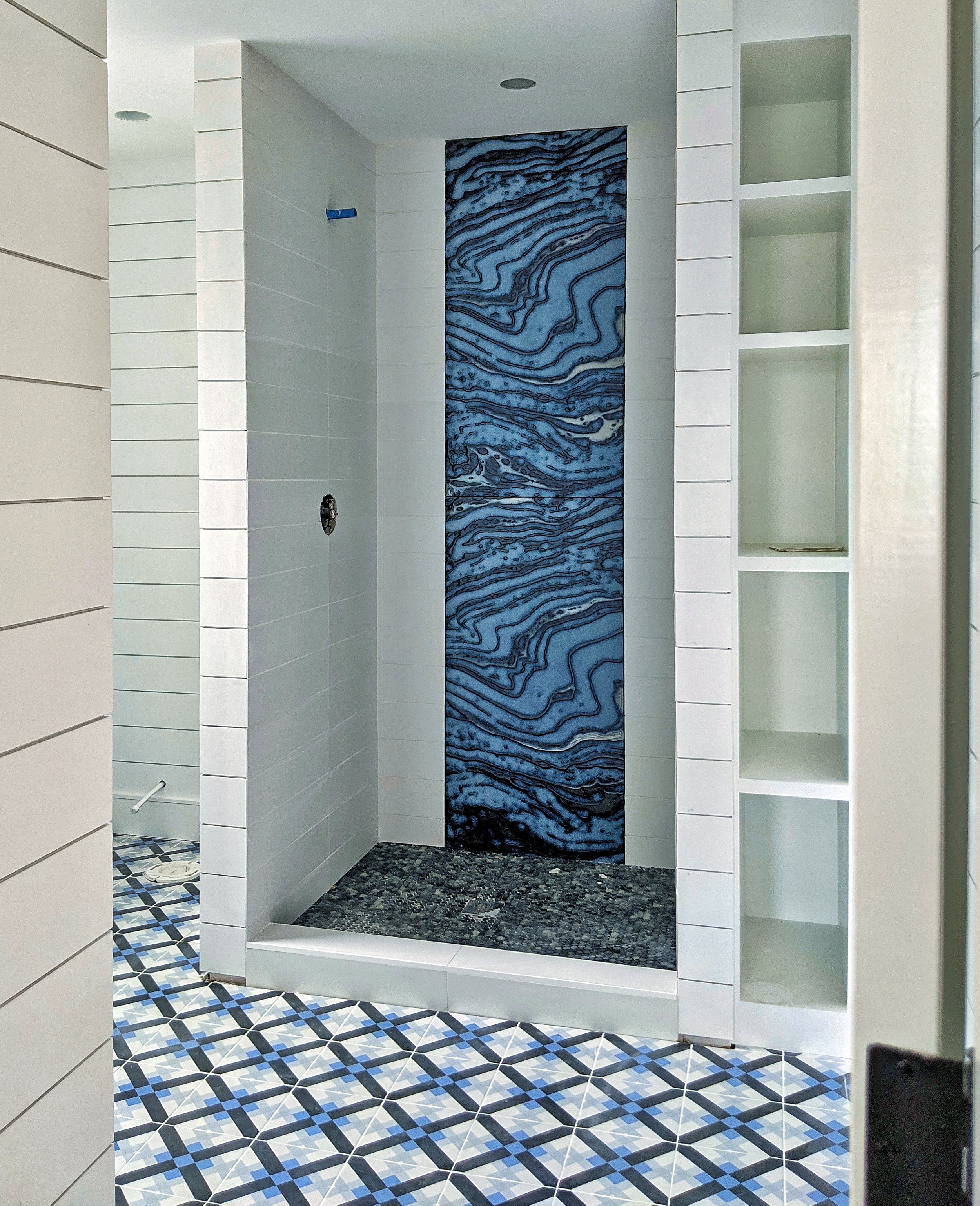 Modern shower with blue and black marbled accent wall, geometric patterned floor tiles, and white tiled walls, featuring built-in shelving on the right.