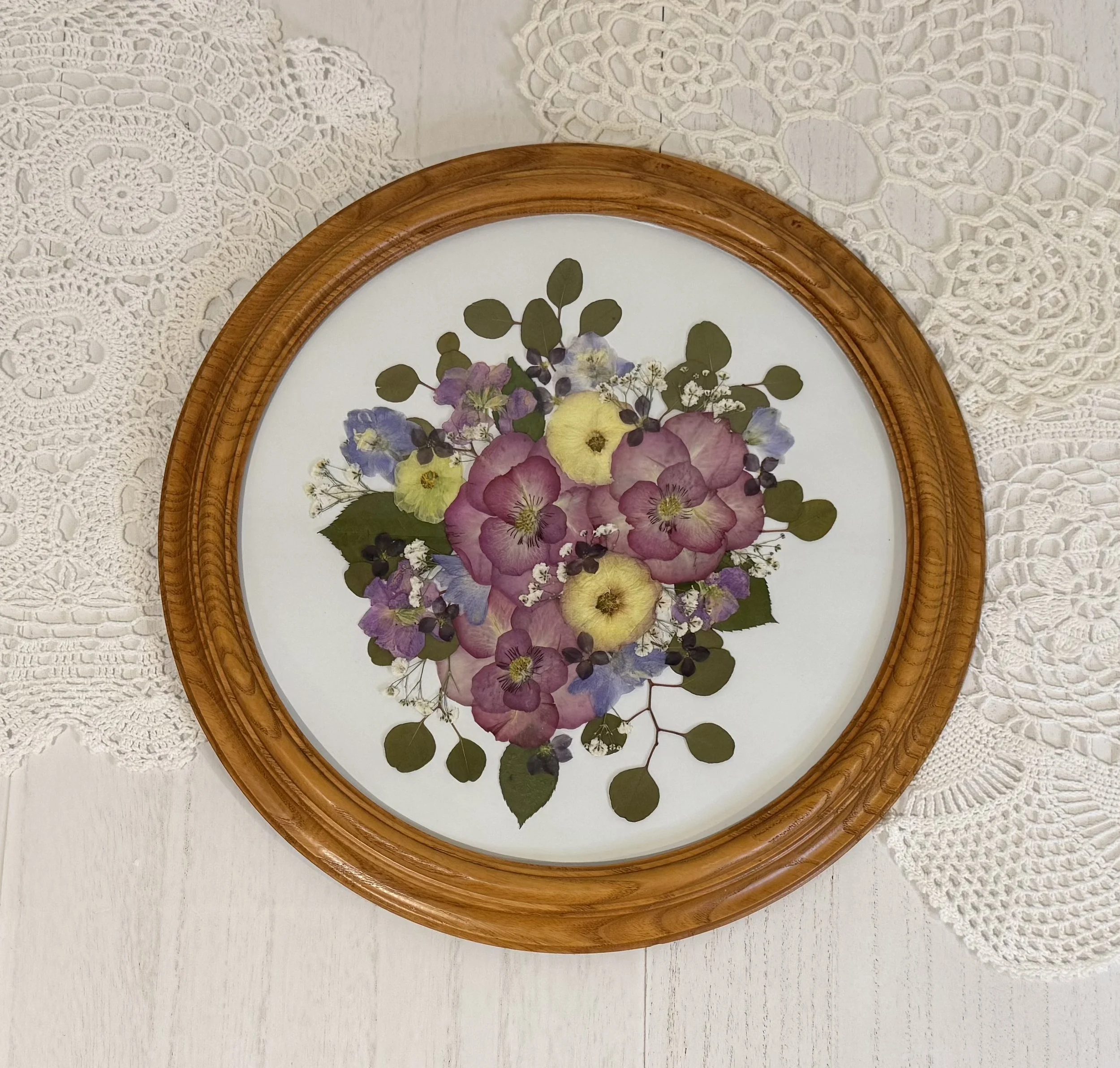 A framed pressed flower art piece with pink, yellow, purple, and white flowers and green leaves on a white background, placed on a white lace tablecloth.