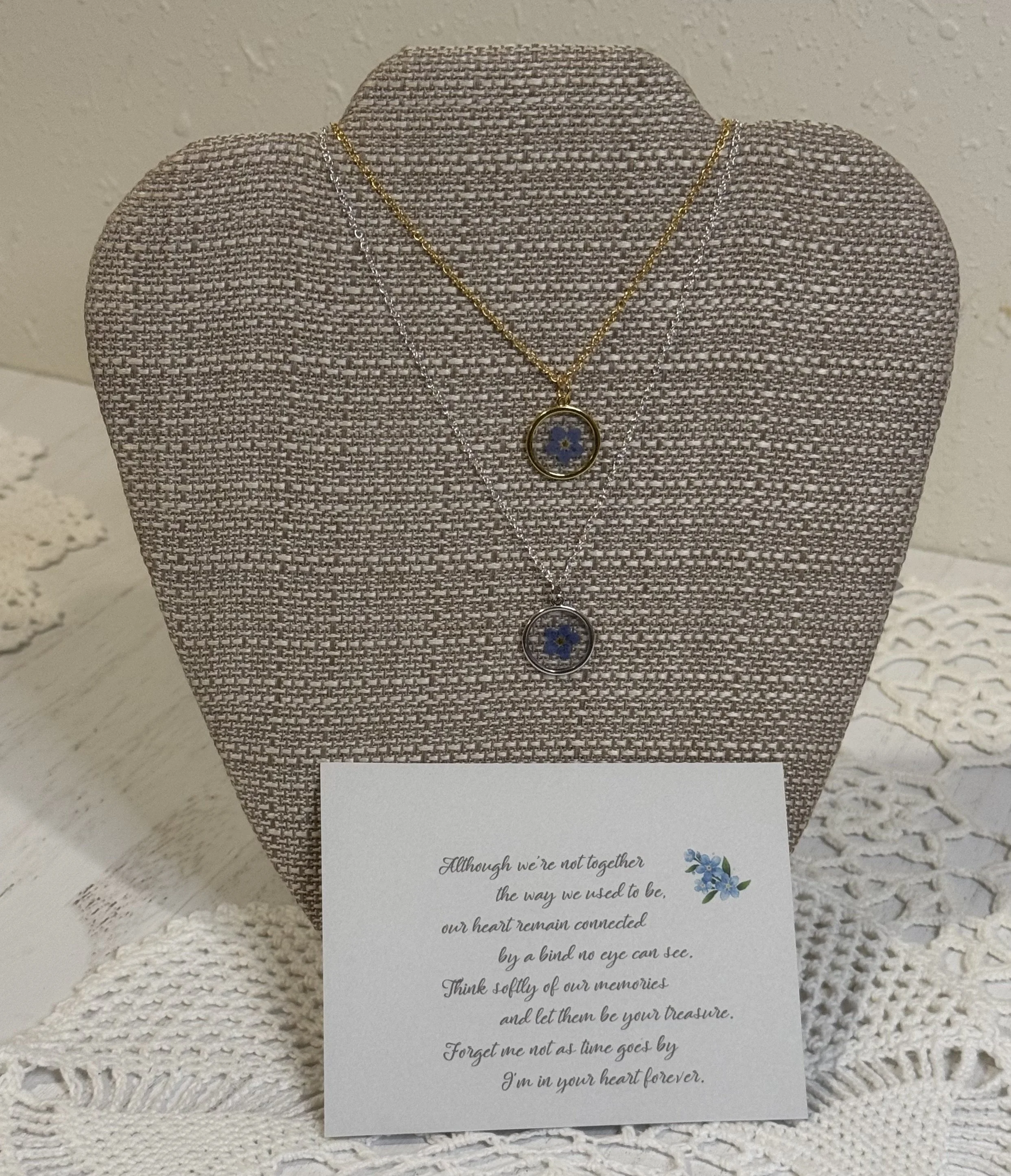 Two layered necklaces with blue flower pendants displayed on a textured fabric bust, with a heartfelt message card underneath.