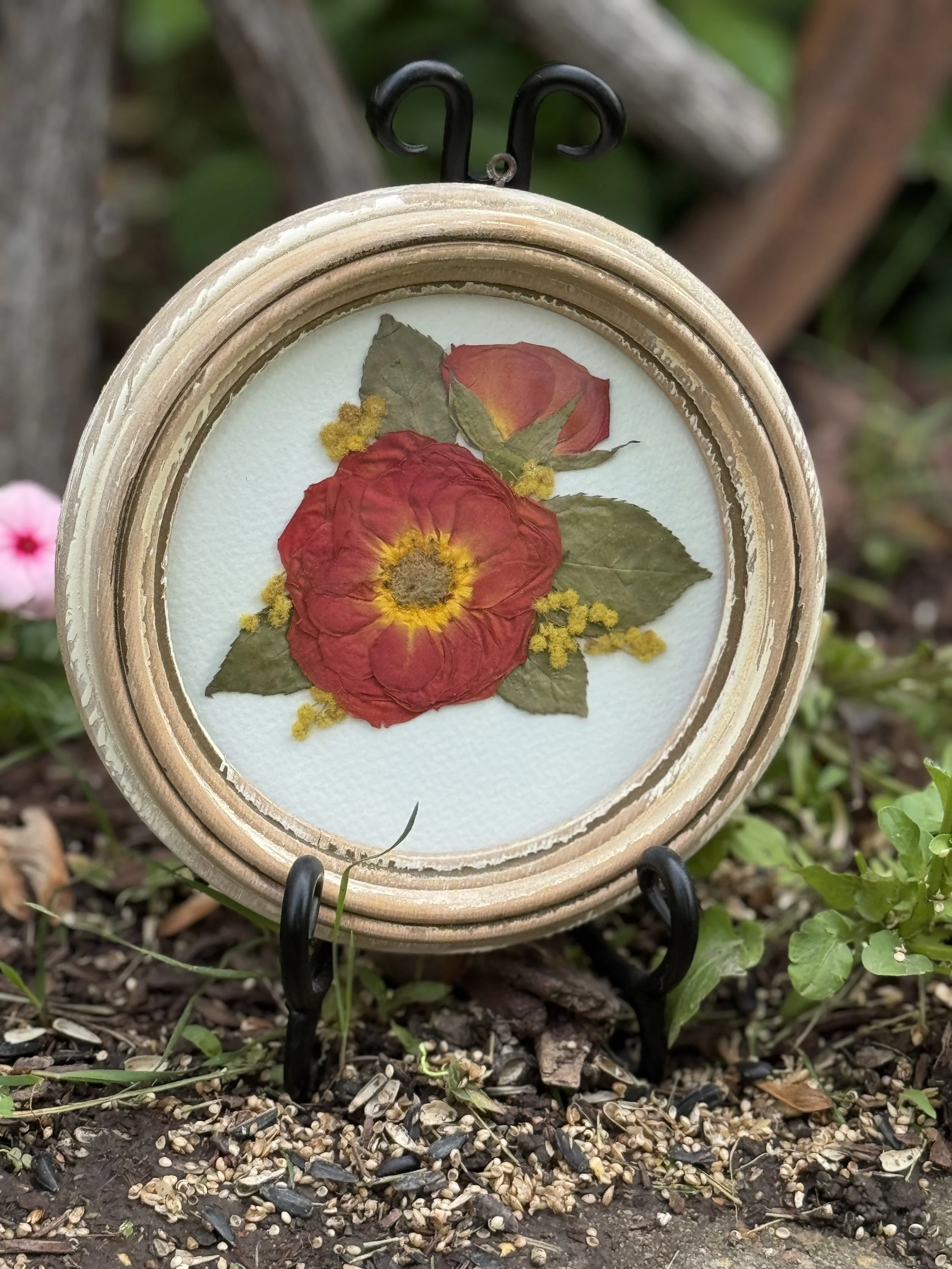 A decorative oval frame with a dried flower arrangement, featuring a red flower, green leaves, and small yellow accents, is displayed outdoors on a small stand.