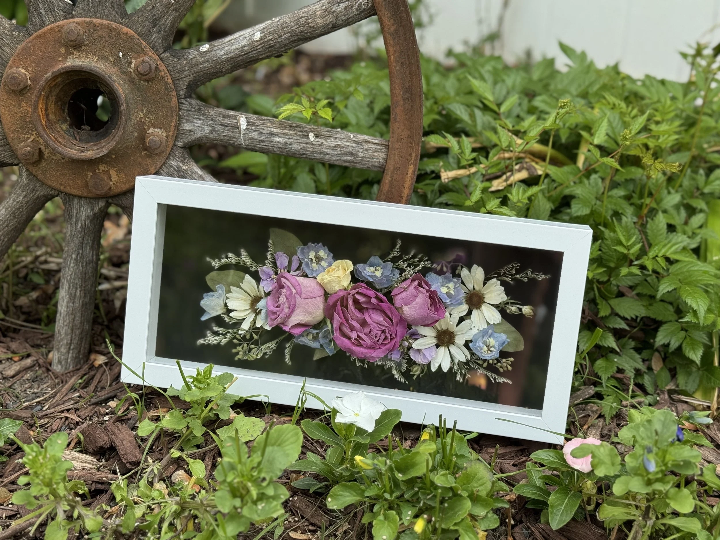 A framed floral artwork featuring pink roses, white daisies, and blue flowers, resting on the ground among green plants and mulch, with a rusty, weathered wagon wheel in the background.