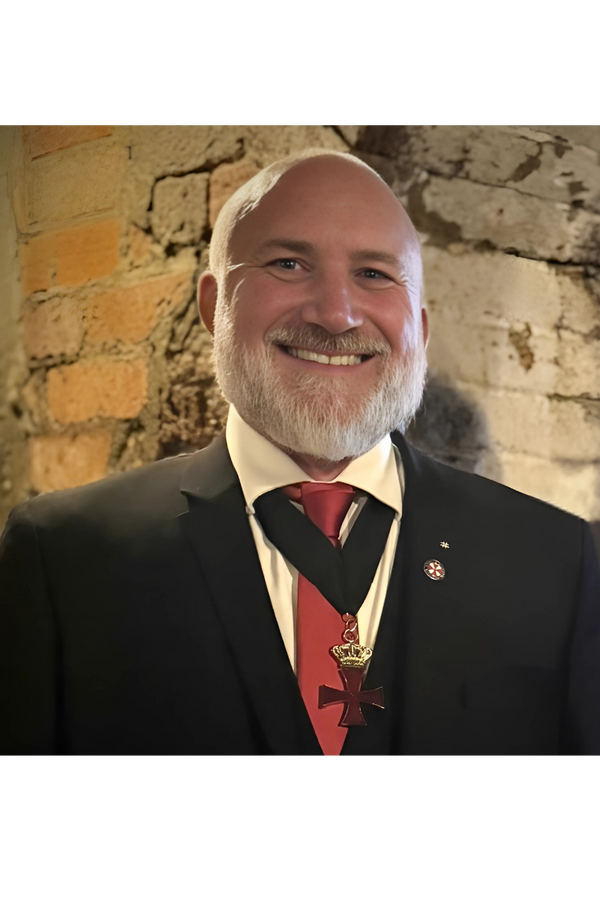A man with a white beard in formal attire, wearing a black suit, white shirt, red tie, and a medal with a cross and crown, smiling in front of a brick and stone wall.