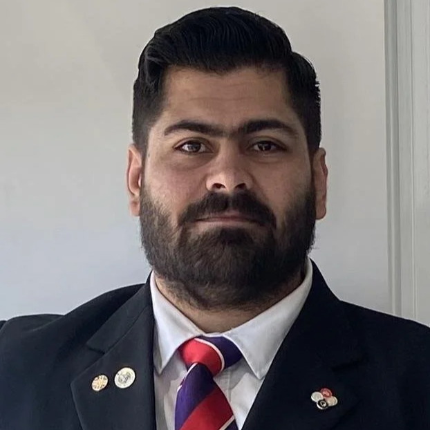 A man in a black suit with a white shirt and red tie standing indoors near a brick wall.