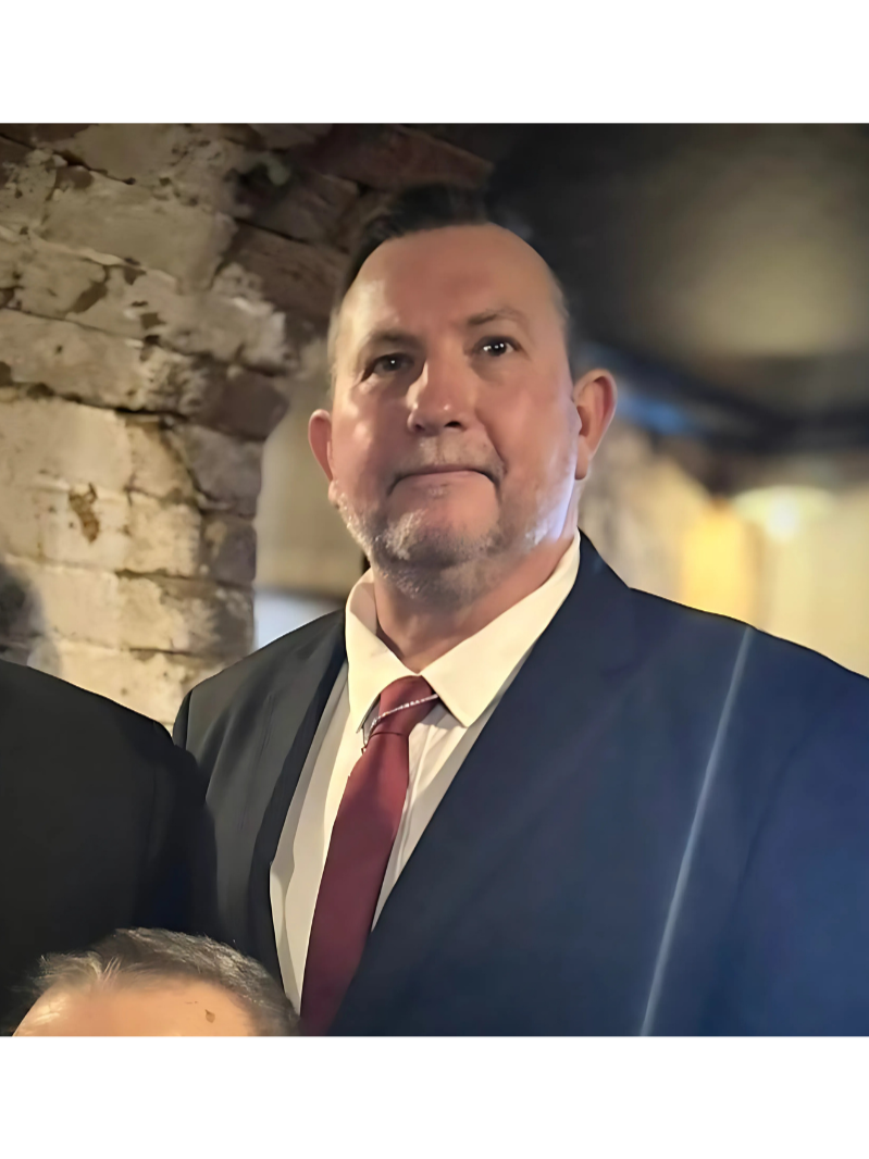 A man in a black suit with a white shirt and red tie standing indoors near a brick wall.