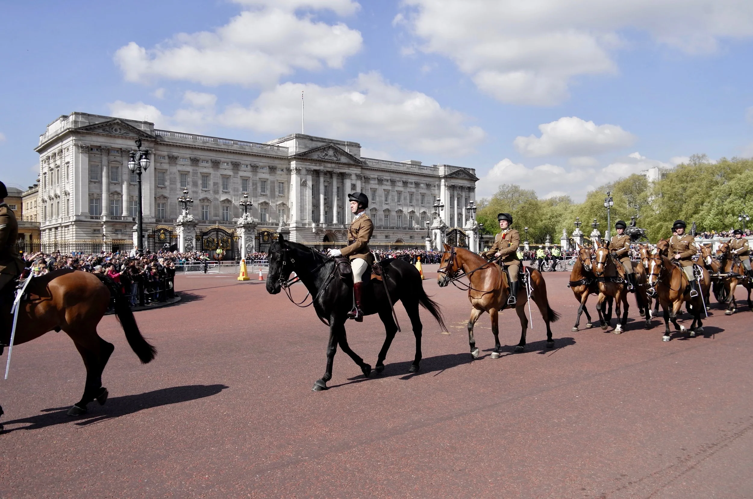 A parade of mounted soldiers in uniform riding horses in front of Buckingham Palace, with a crowd of spectators behind barriers on a sunny day.