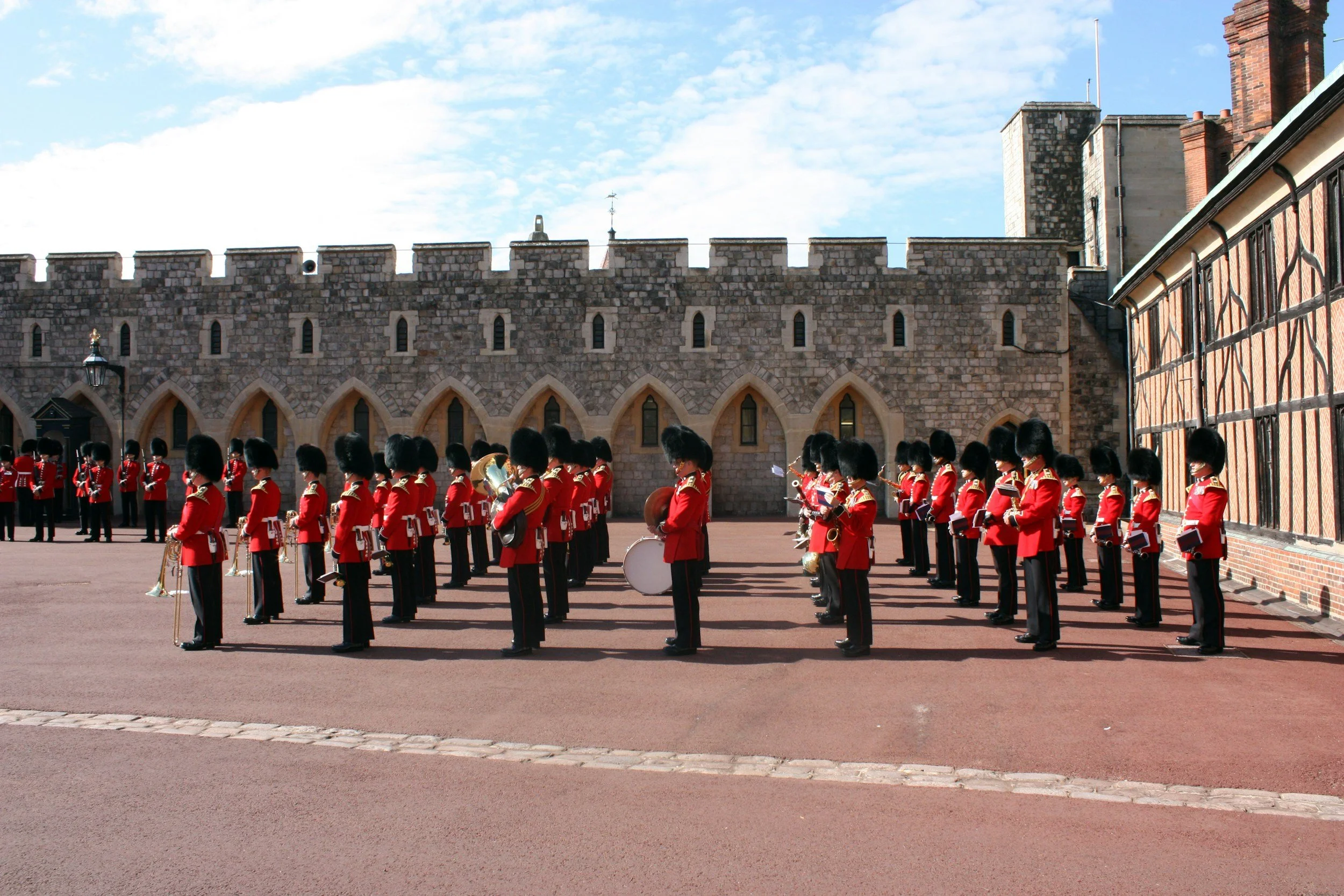 A group of uniformed soldiers standing in formation outdoors in front of a stone castle wall, with some holding musical instruments.