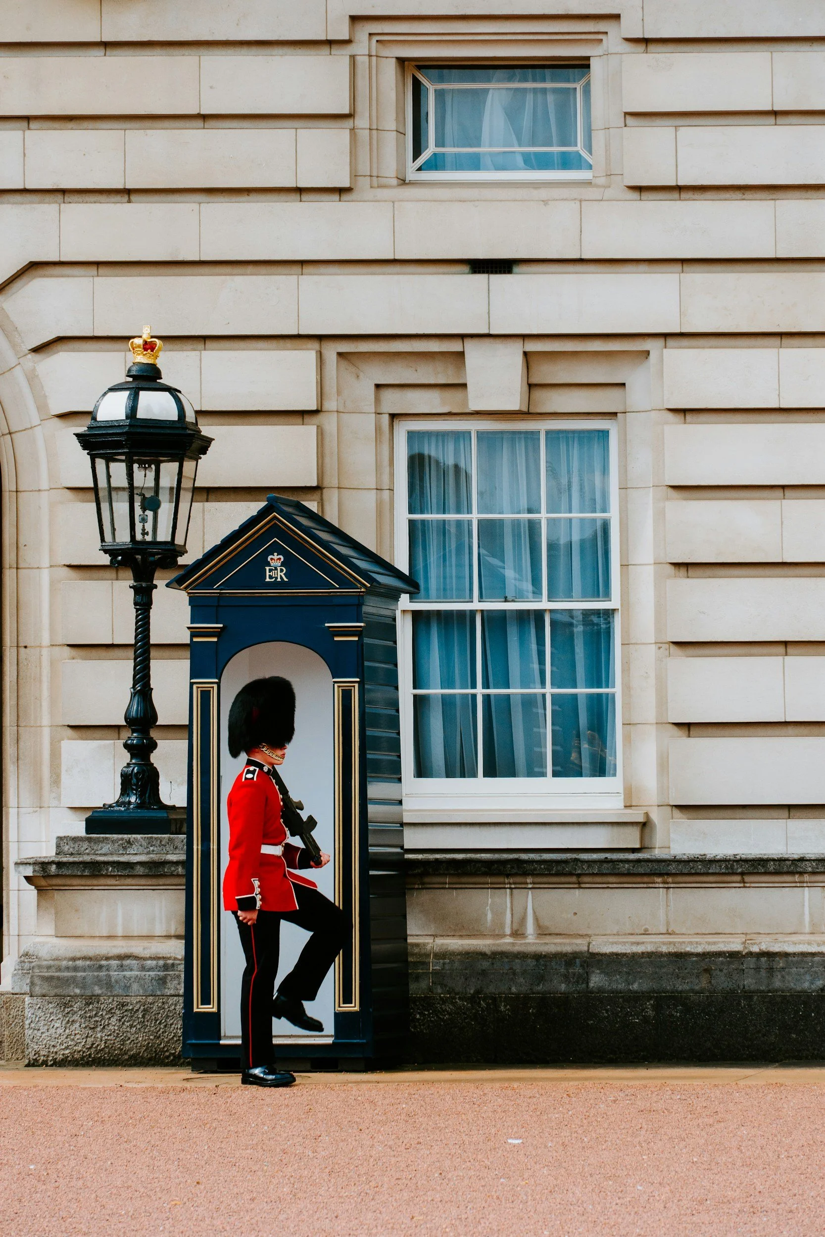 A Royal guardsman in red uniform and tall black hat stands inside a blue sentry box with gold trim in front of a beige stone building. A black lantern post with a small crown on top is beside the sentry box.