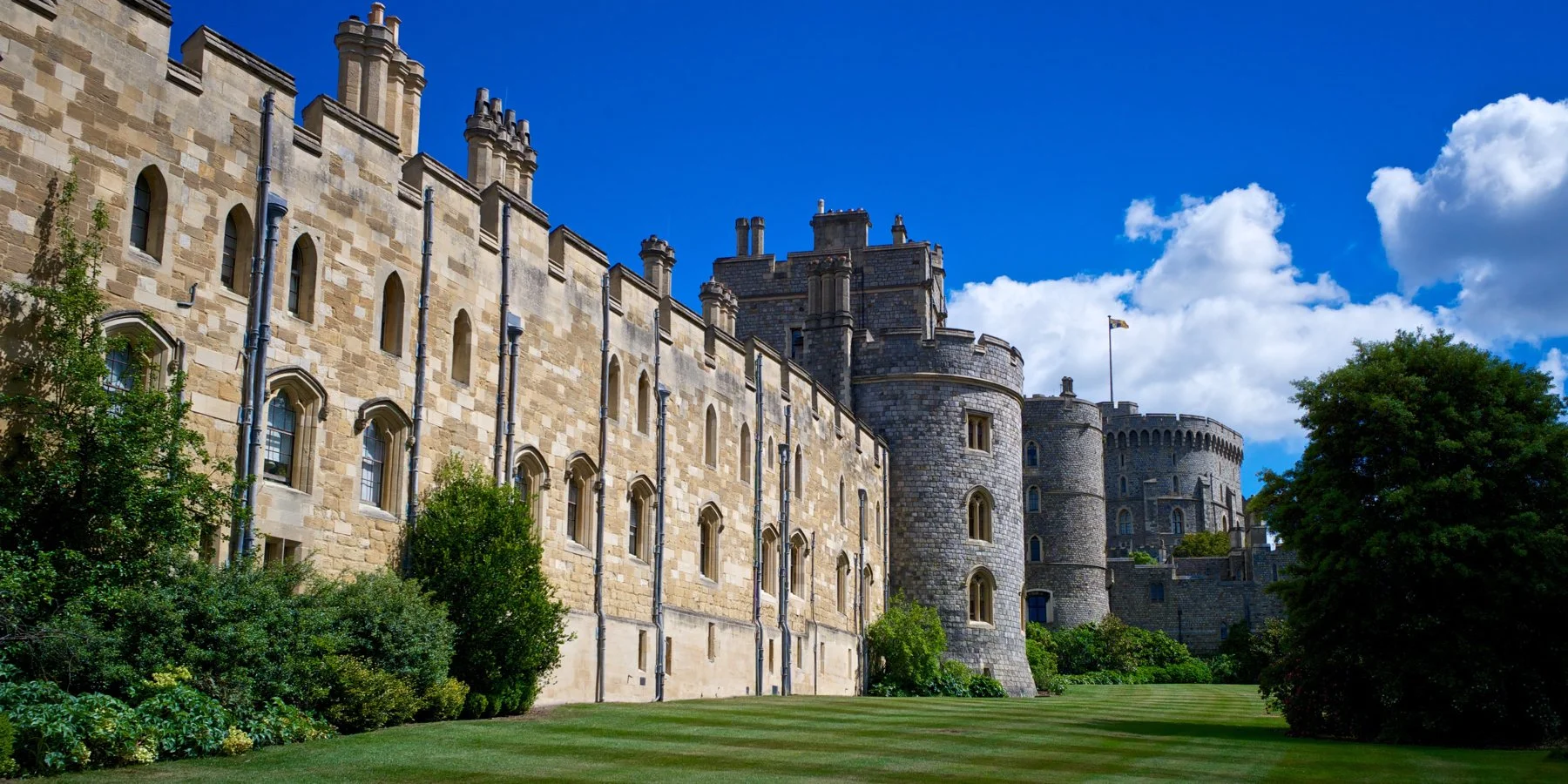 A large stone Windsor Castle with turrets, surrounded by green grass and bushes, under a partly cloudy blue sky with some white clouds.