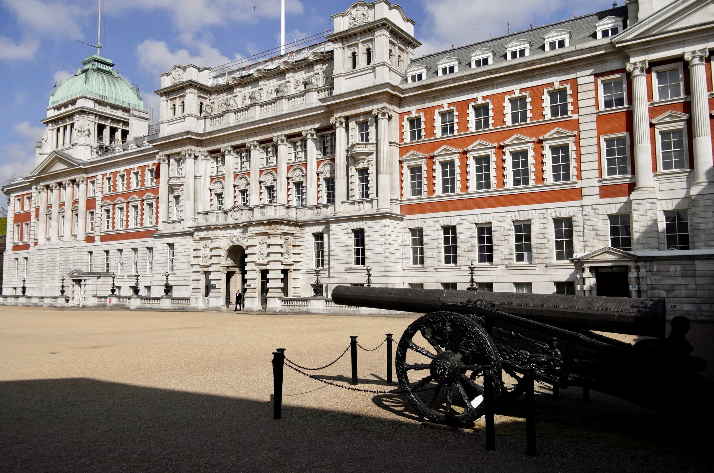 Large historic building with stone and red brick exterior, multiple windows, decorative columns, and a gold-domed tower. A black cannon is in the foreground on a gravel area. The building appears to be a government or royal residence.