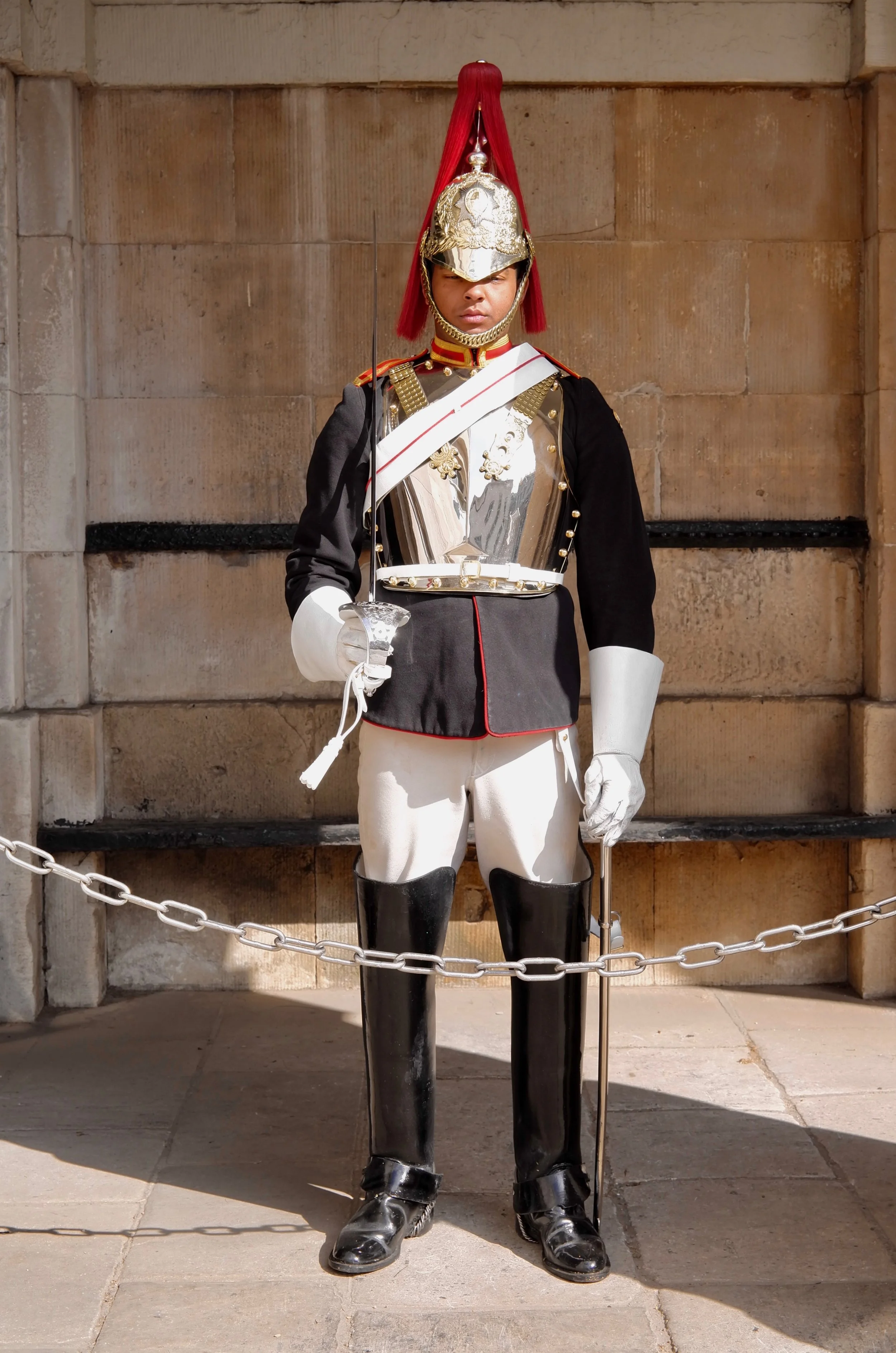 Soldier in ceremonial uniform standing guard with a rifle, wearing a silver helmet with a red plume, black coat with gold accents, white trousers, and black boots.