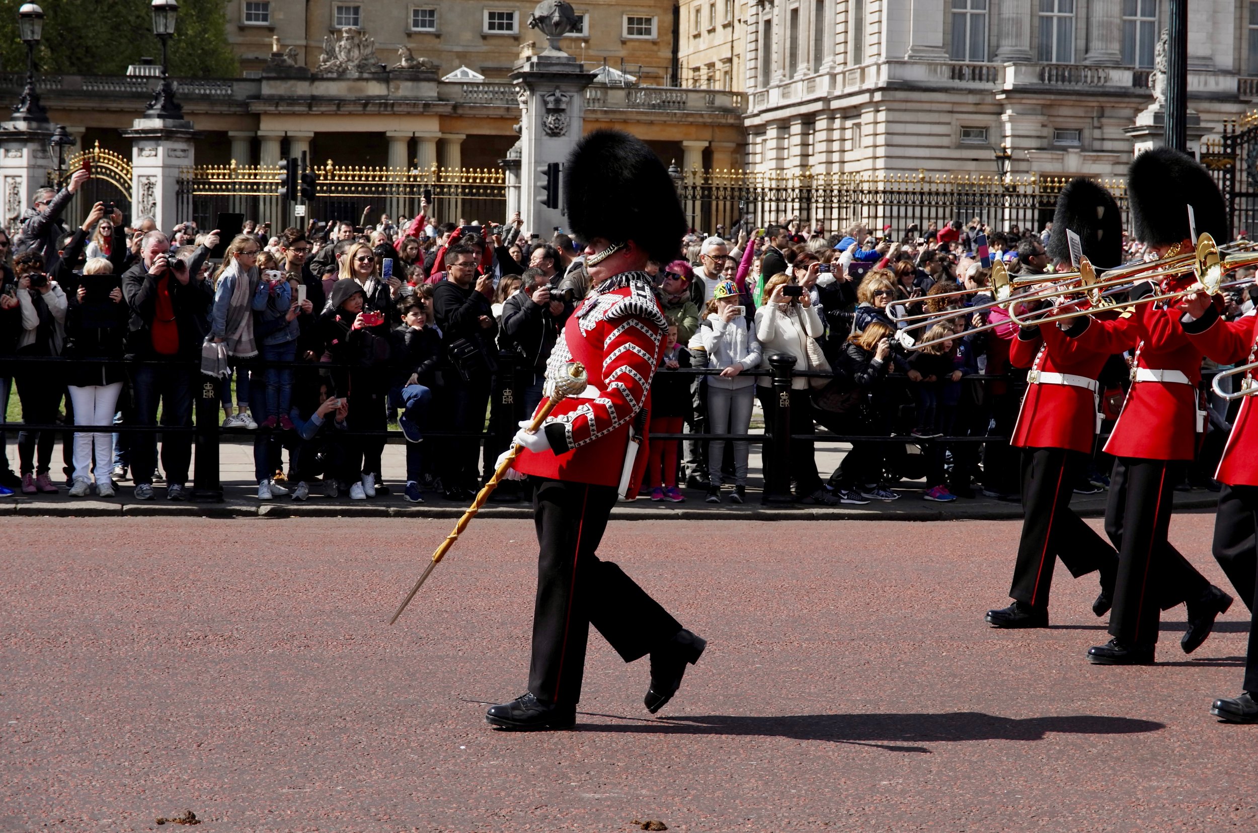 Queen's Guard soldiers in red uniforms and bearskin hats marching during a parade outside a historic building, with a crowd of spectators taking photos behind a black fence.