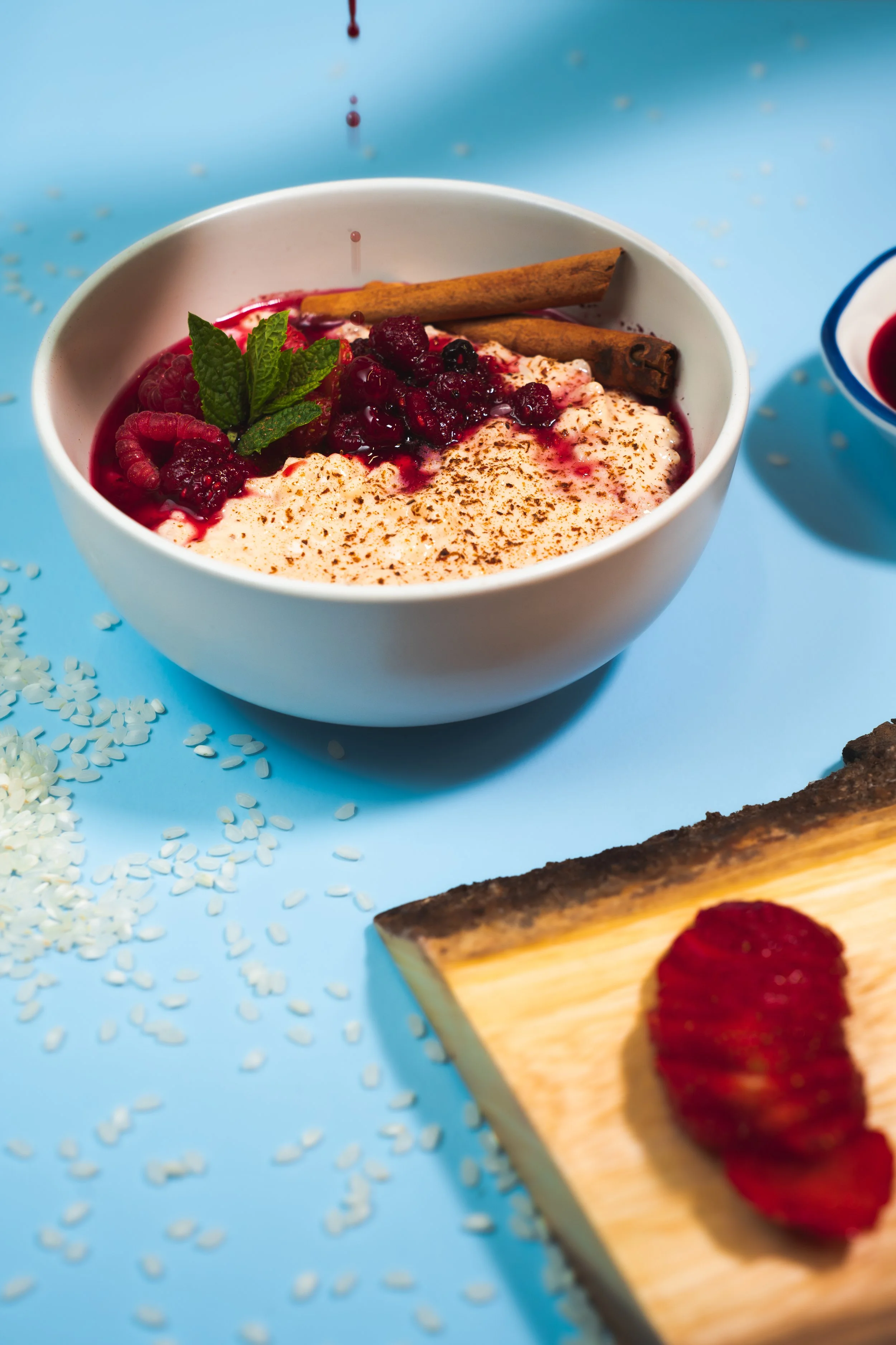 A bowl of dessert garnished with fresh berries, mint, and a cinnamon stick, with some spices and a wooden tray with a strawberry nearby.