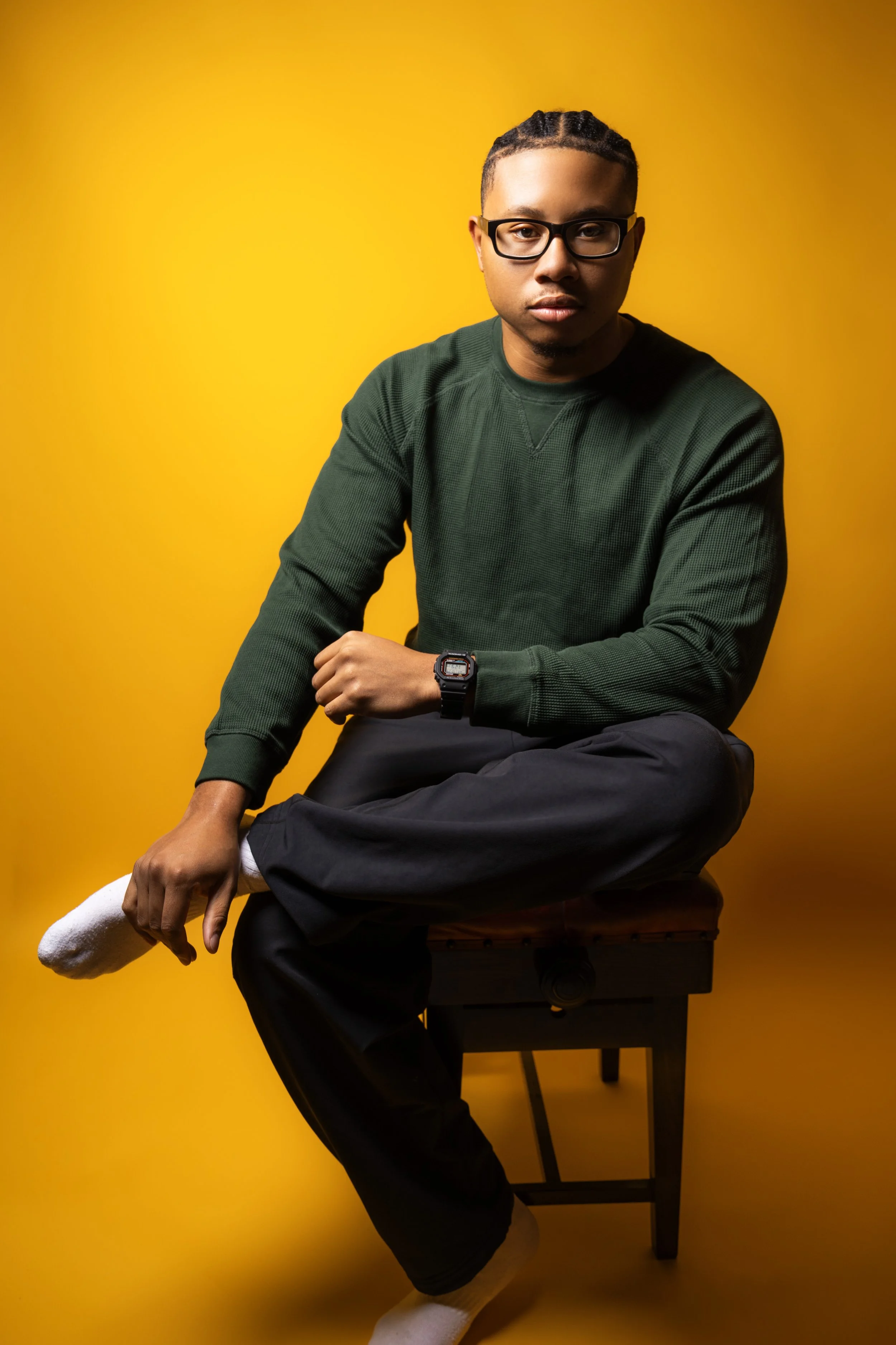 A young man with glasses wearing a green sweater and black pants, sitting cross-legged on a wooden stool against a yellow background, holding one shoe with white socks.