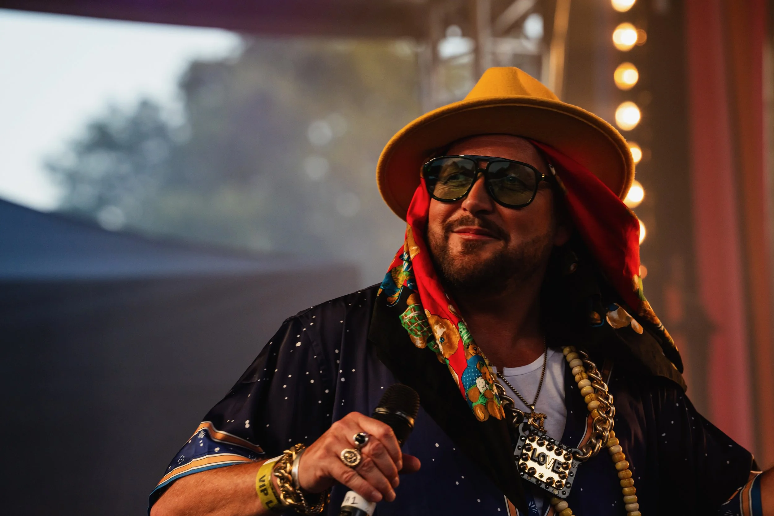 A man wearing a yellow wide-brimmed hat, sunglasses, a red bandana, and colorful jewelry, holding a microphone on stage.