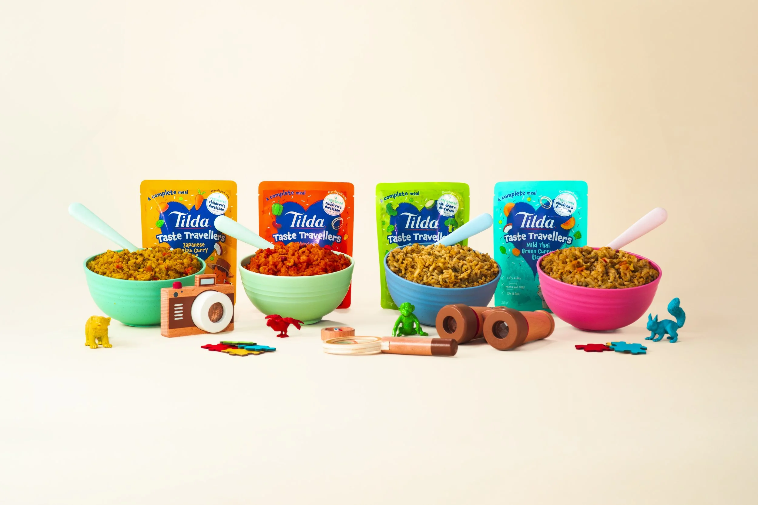 Four colourful bowls of Tilda Taste Travellers packaging behind each, along with miniature animals, camera, and small wooden toy tools on a light-coloured background.