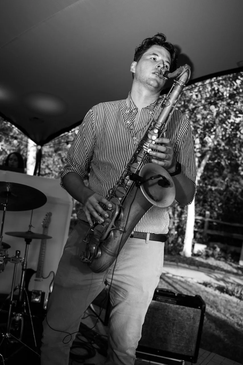 A man playing the saxophone during an outdoor performance, with a drum set and electric guitar in the background.