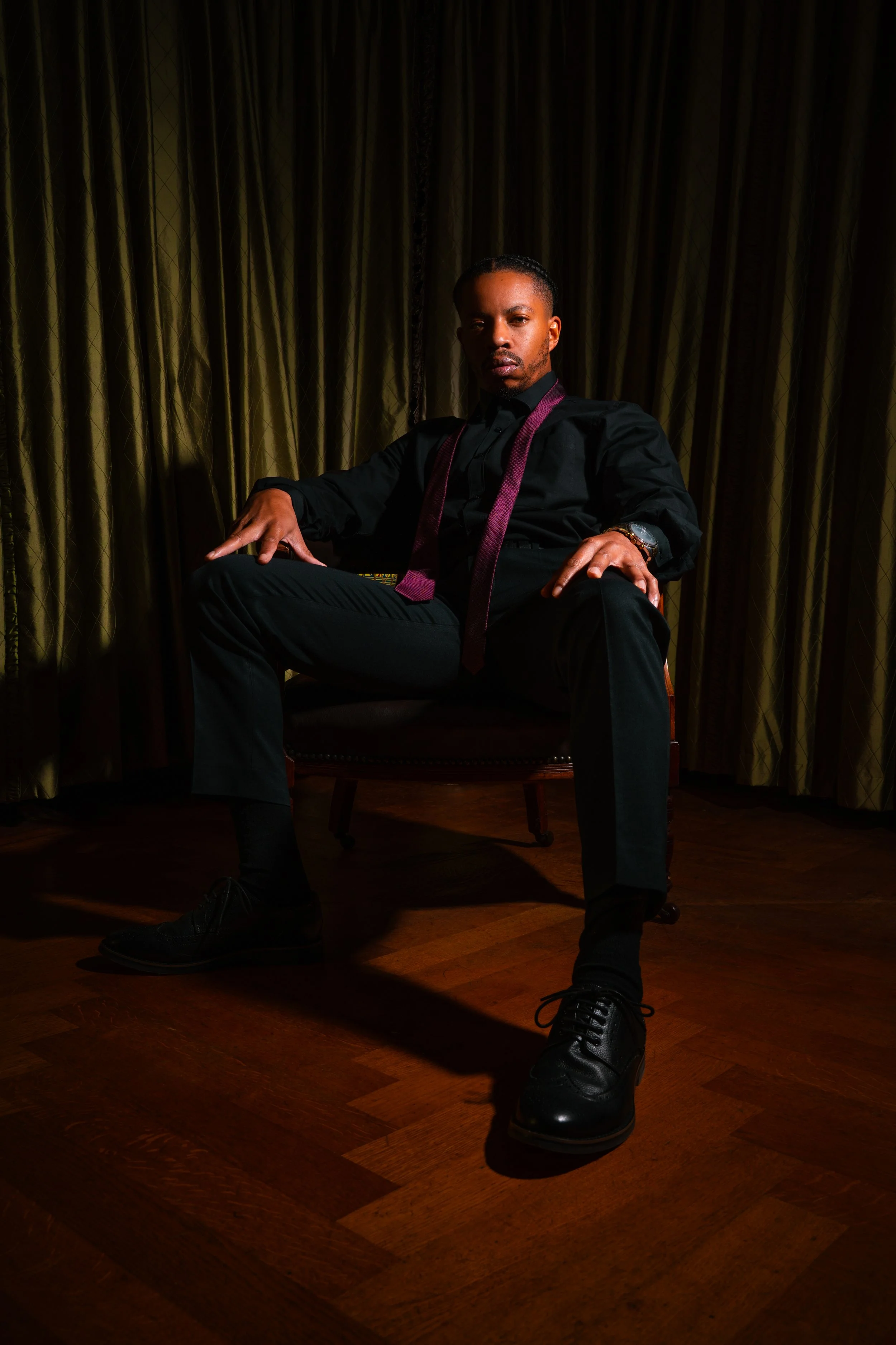 A man sitting confidently in a chair in front of a dark curtain, wearing a black shirt, trousers, shoes, and a purple tie with a watch on his wrist.
