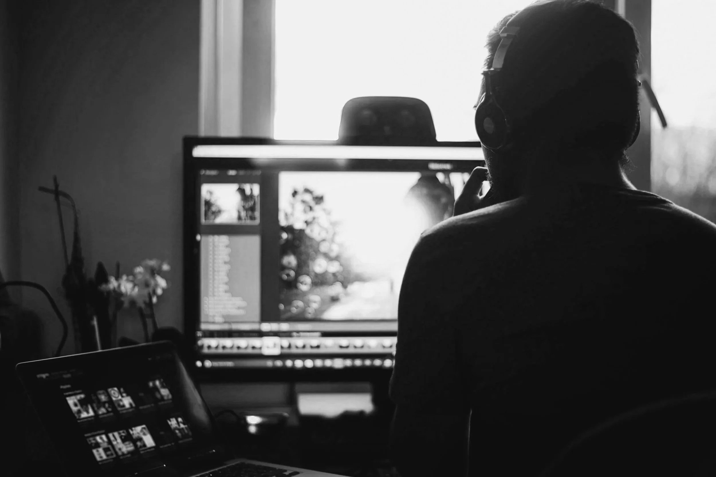 Silhouette of a person wearing headphones working on a computer with photo editing software, with a tablet nearby showing a grid of images, in a dimly lit room with light coming through a window.