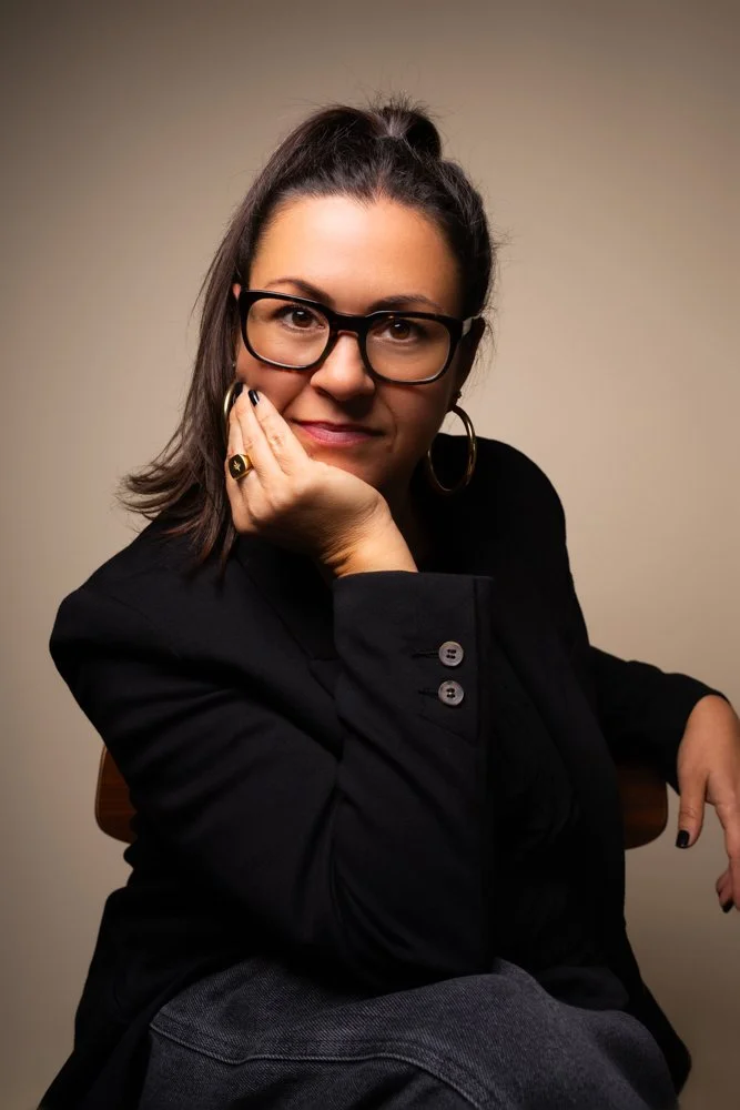 A woman with dark hair in a ponytail, wearing glasses, a black blazer, and hoop earrings, sitting against a plain beige background.