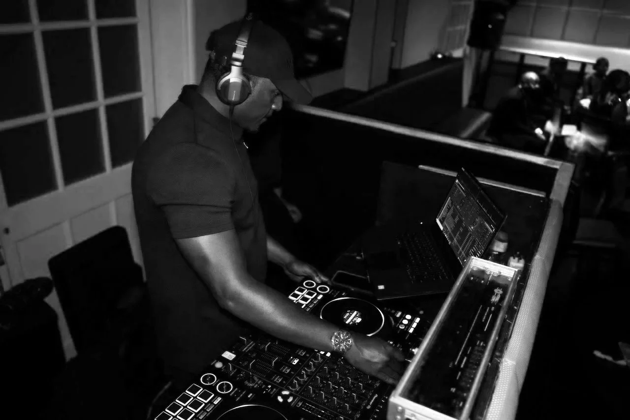 A DJ wearing a cap and headphones operating DJ equipment including a laptop, mixer, and turntables in a club setting.