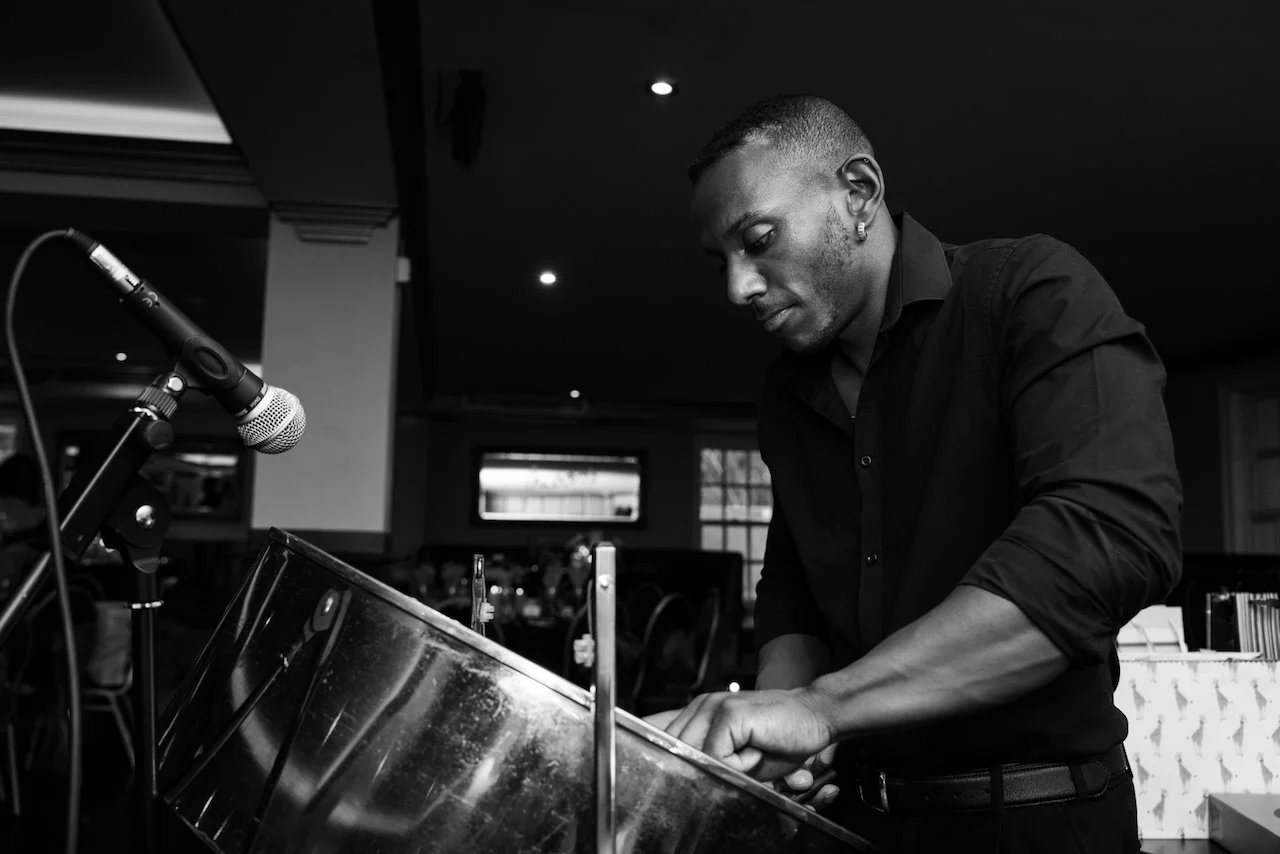 A man in a black shirt plays a steel drum with a microphone set up nearby in a dimly lit indoor setting.