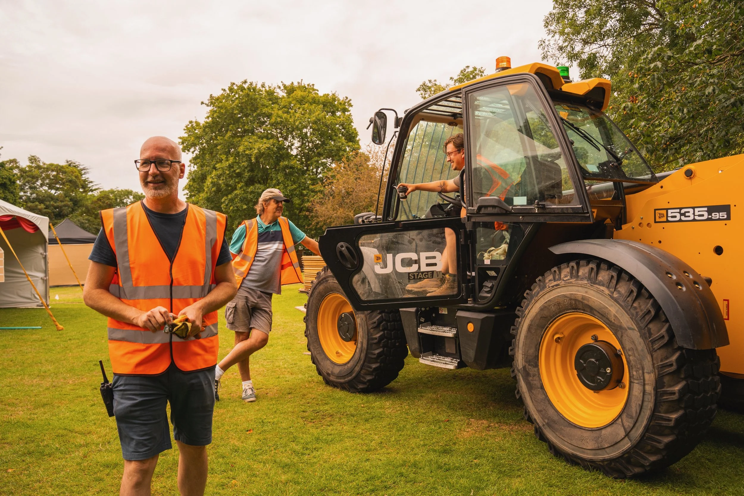 Three men in safety vests on a grassy field, with a large yellow construction vehicle and tents in the background. One man is seated in the vehicle, smiling, while the other two stand nearby, talking and smiling.