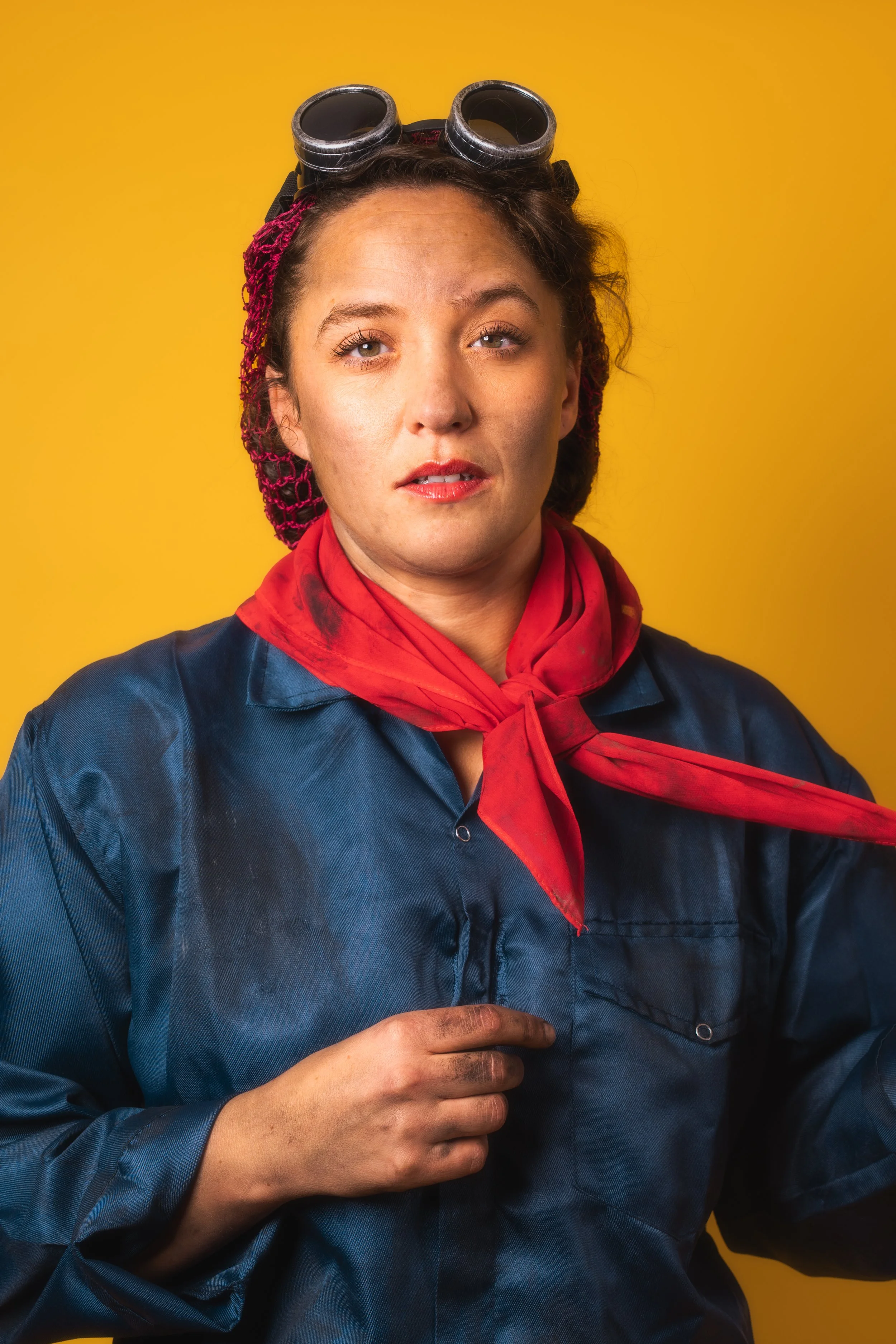 A woman dressed in a Rosie the Riveter-style throwback costume, wearing a blue work shirt, red bandana, red neckerchief, and work goggles on her head, against a yellow background.
