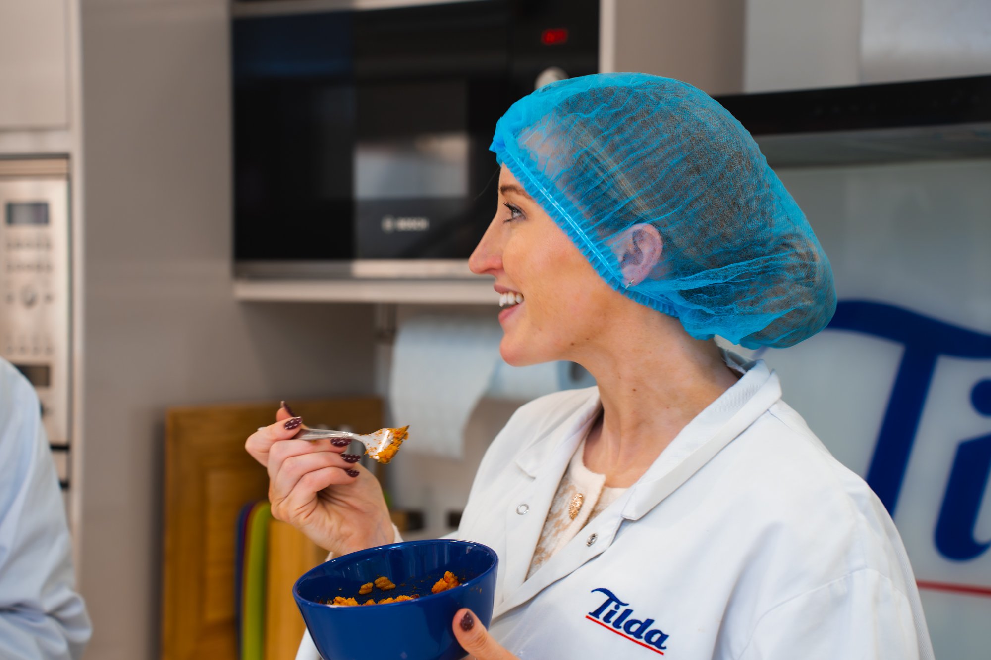 Woman in white chef's coat and blue hairnet tasting food while holding a blue bowl in a commercial kitchen.