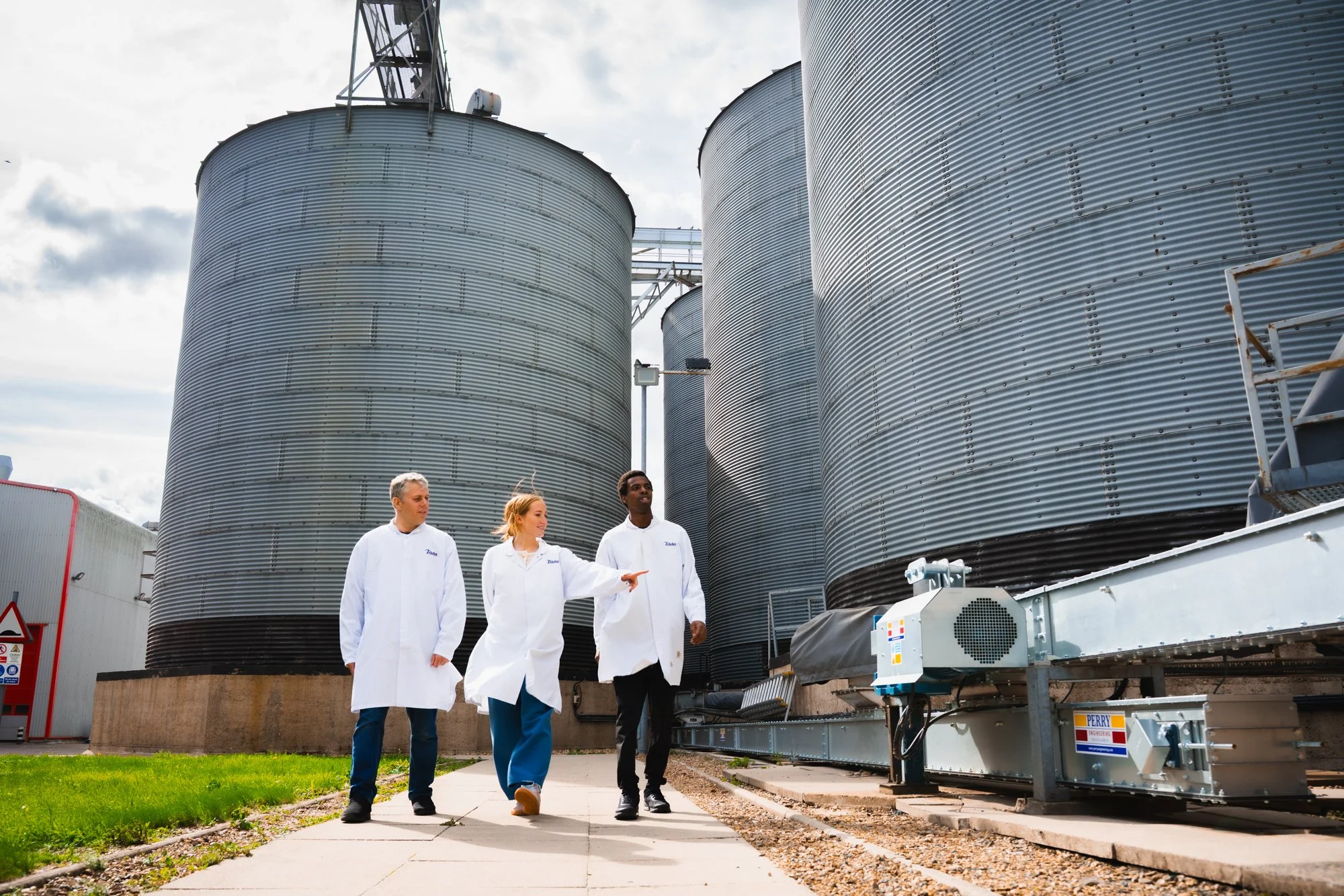 Three scientists in lab coats walking outside industrial storage tanks, discussing or inspecting the site.