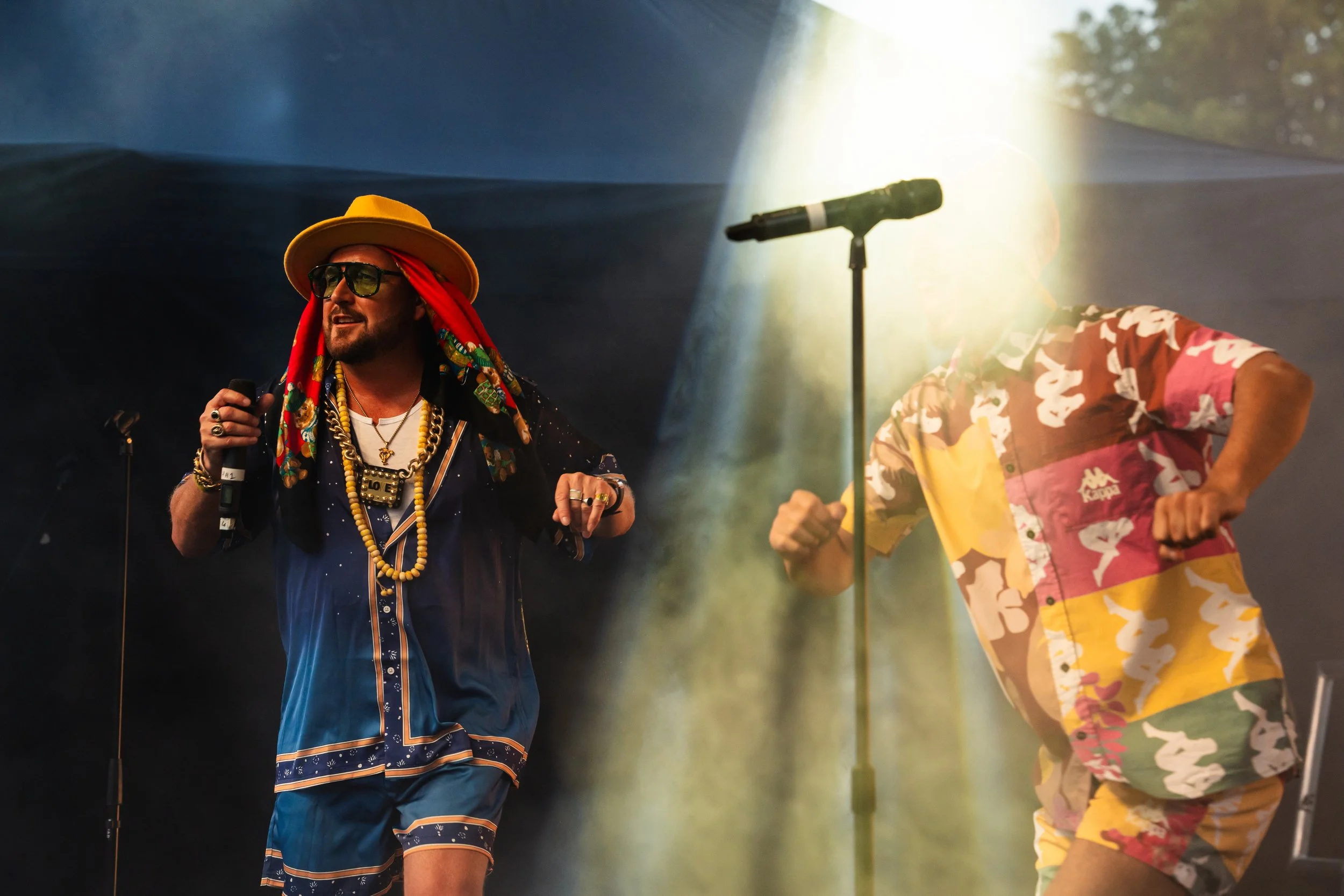 Two men performing on stage at a concert, both dressed in colorful, tropical-themed clothing. One man is wearing a large yellow hat, sunglasses, and various accessories, holding a microphone. The other man is wearing a multicolored shirt with a camouflage pattern and sunglasses, with his head down near a microphone stand. Bright stage lights shine behind them.