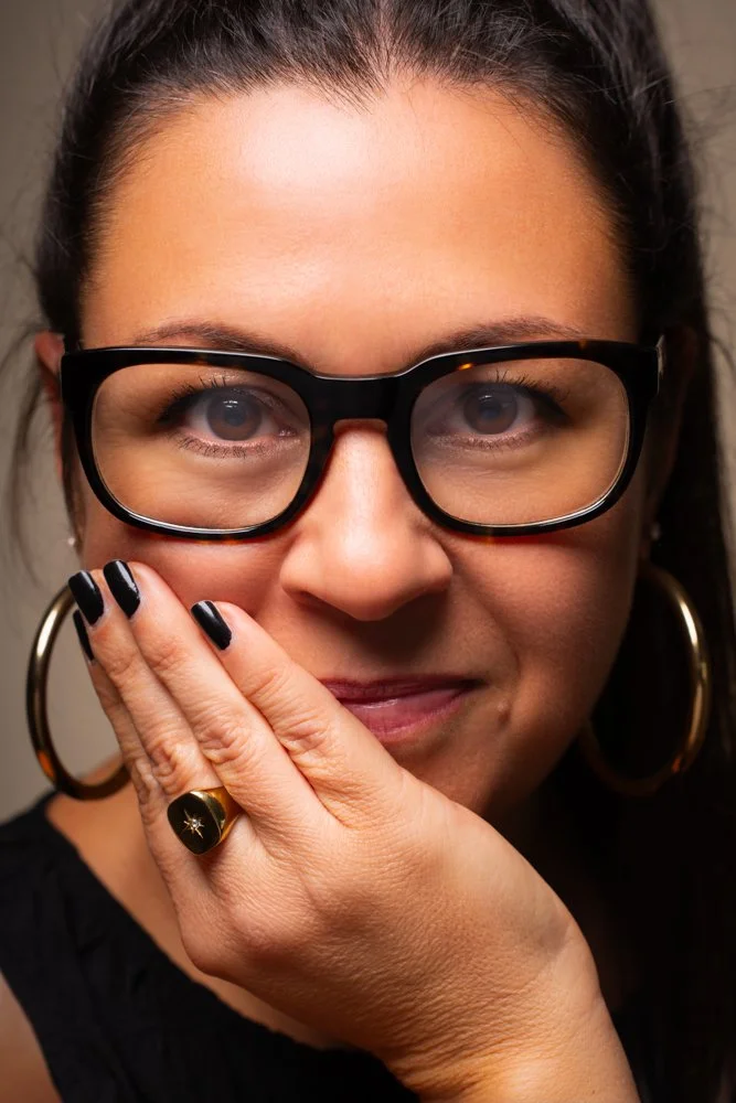 Close-up of a woman with dark hair in a ponytail, wearing large black-rimmed glasses, gold hoop earrings, and a black top. She has her hand near her face, showing a gold ring with a black star and black nail polish. She is smiling subtly.