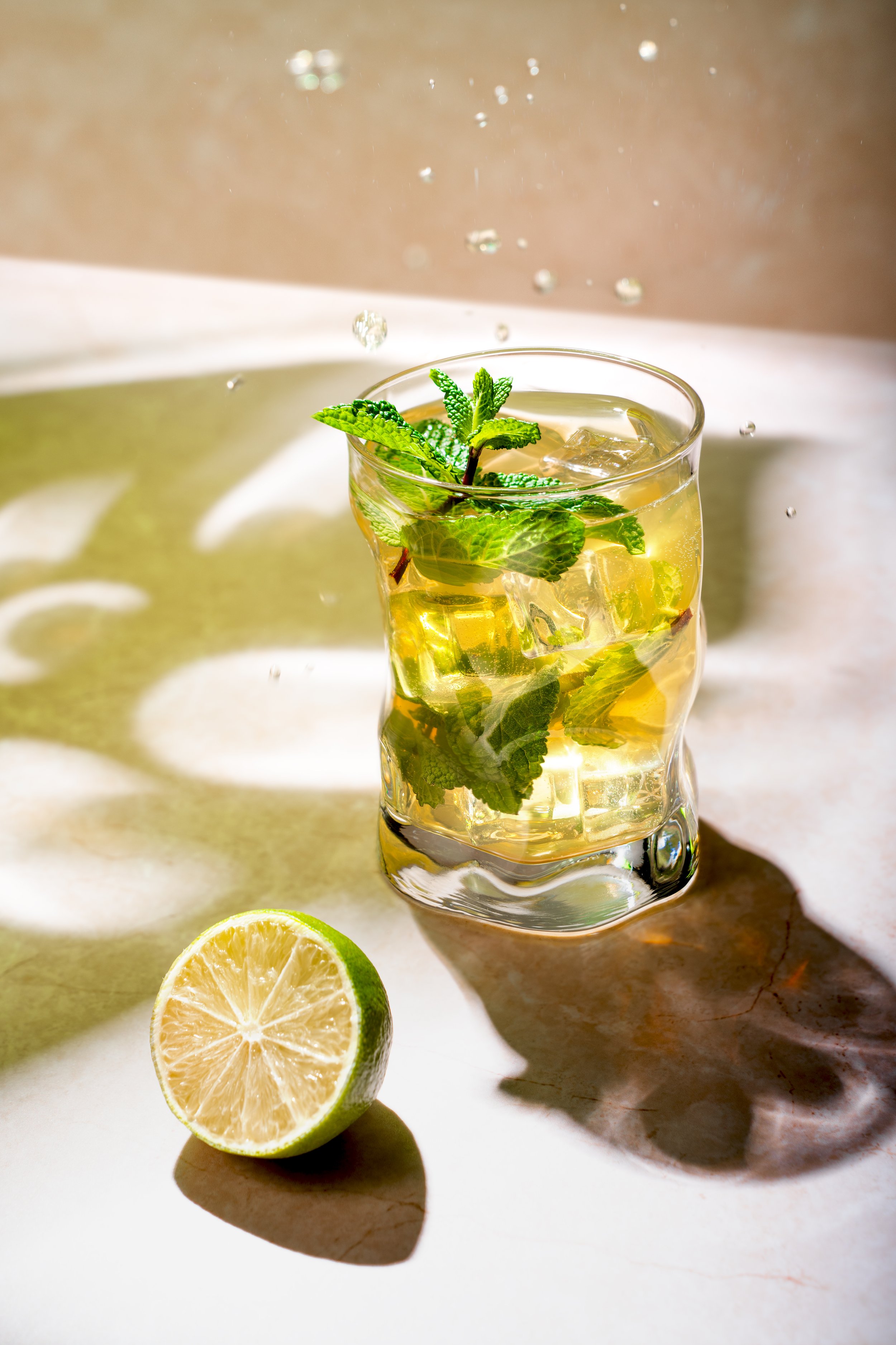 A glass of mojito with mint leaves, lime, and ice on a surface with a sliced lime nearby, with reflections and shadows.