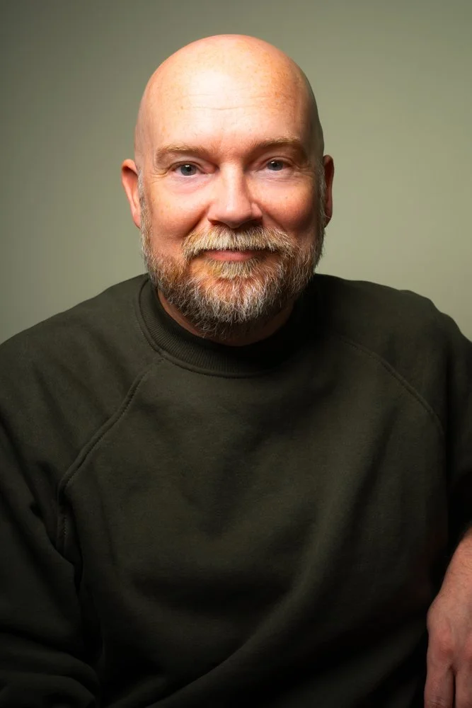 A middle-aged man with a bald head and a beard, wearing a dark sweatshirt, smiling at the camera against a plain, muted background.