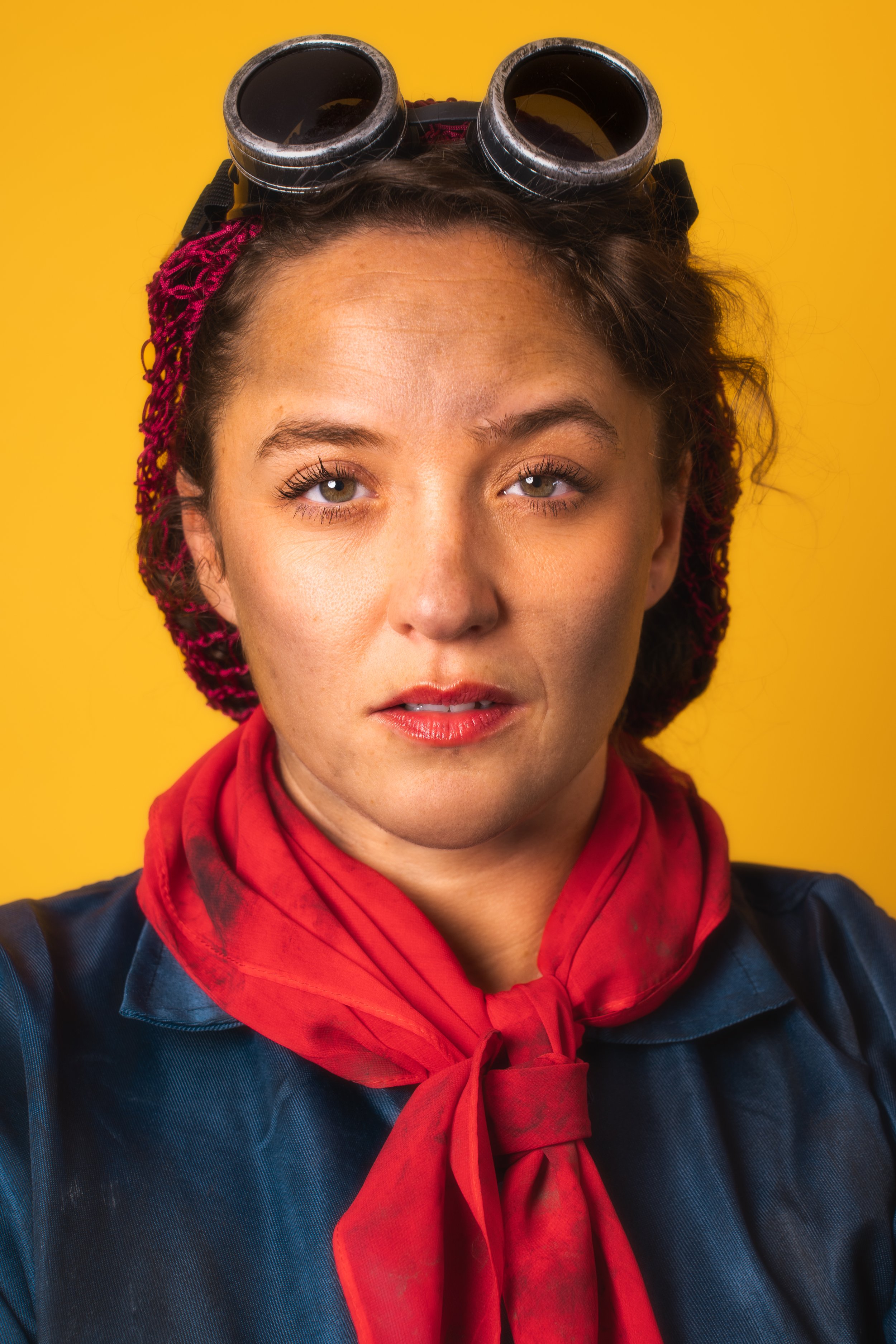 A woman with curly hair, wearing goggles on her forehead, a red scarf around her neck, and a dark jacket against a yellow background.