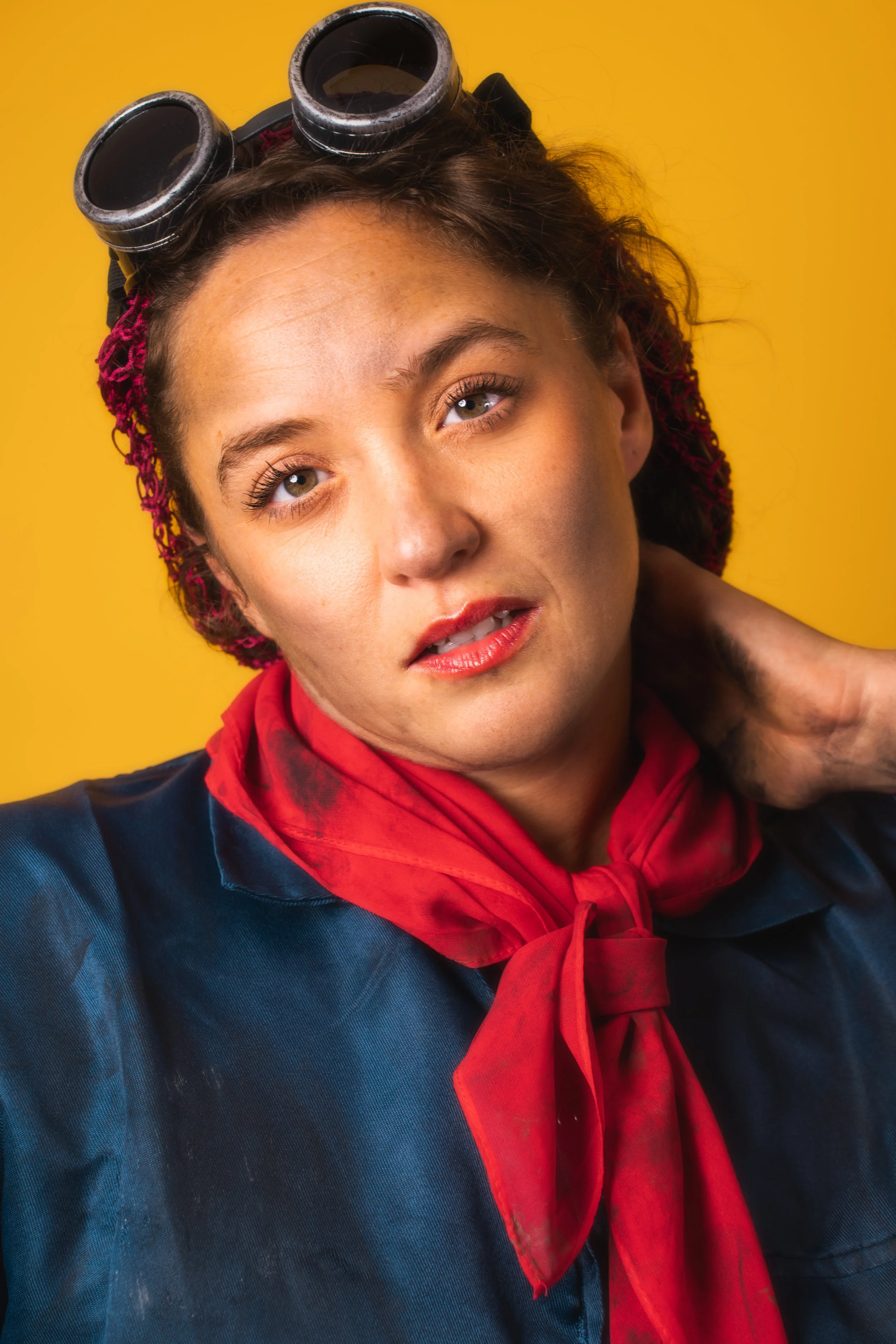 Close-up portrait of a woman wearing goggles on her head, a red scarf, and a dark jacket against a yellow background.
