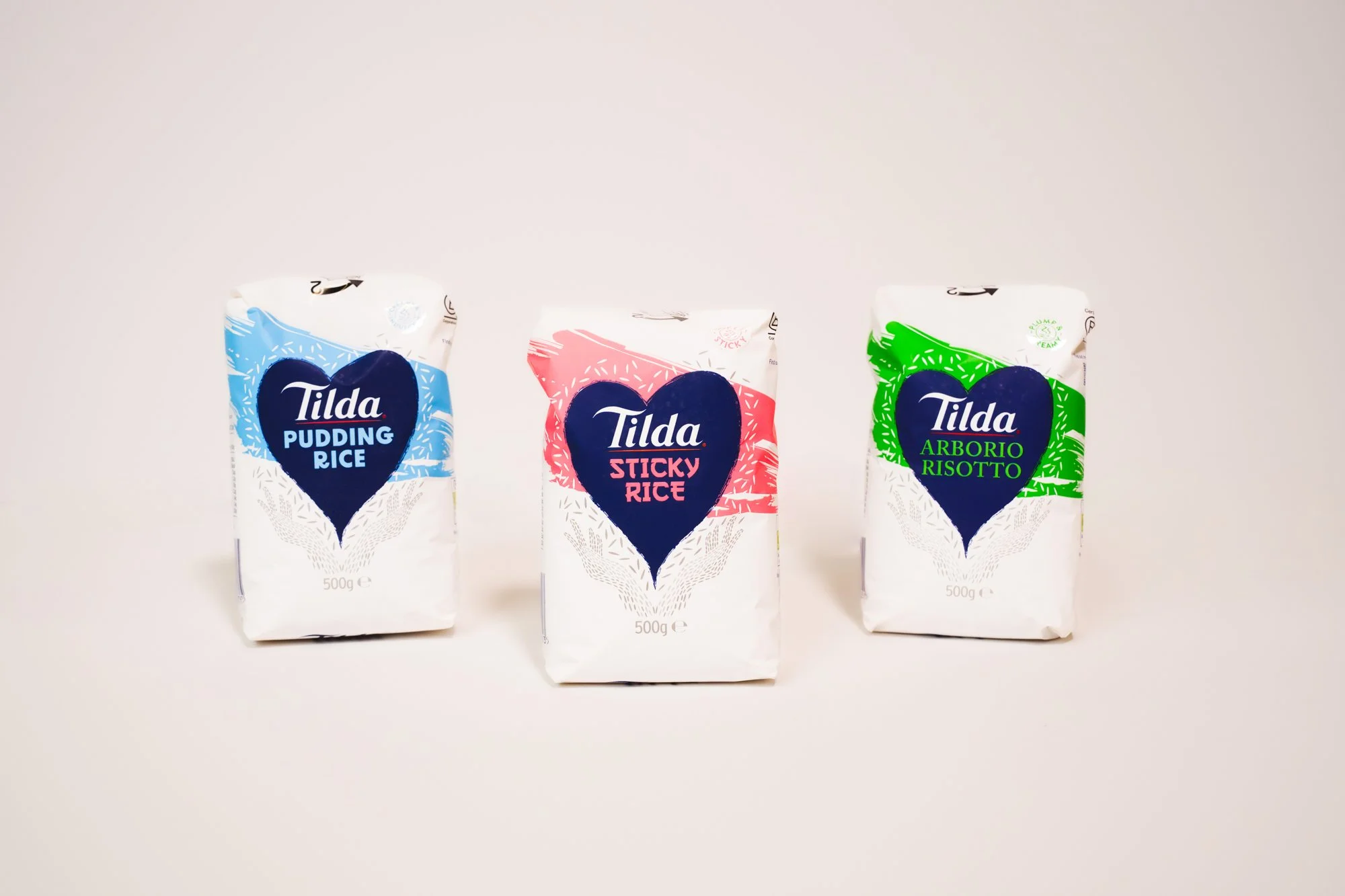 Three bags of Tilda rice, with flavors Pudding Rice, Sticky Rice, and Arborio Risotto, are displayed against a plain background.