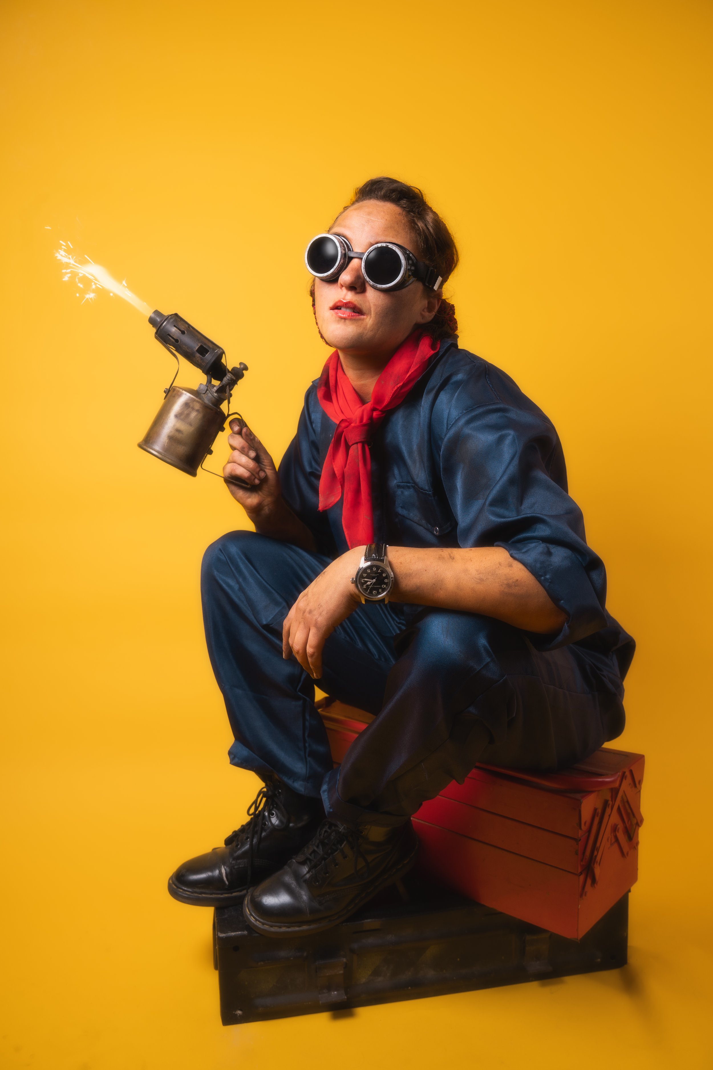 Creative portrait of a woman sitting on a toolbox against a yellow background holding a welding torch