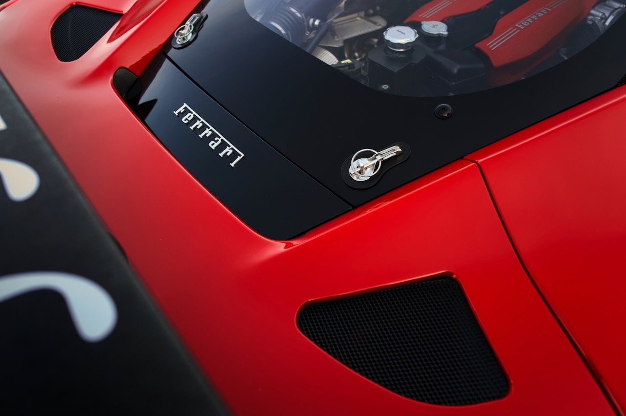 Close-up of a red Ferrari sports car with a black air vent, a windshield with metal latches, and a visible engine with red components.