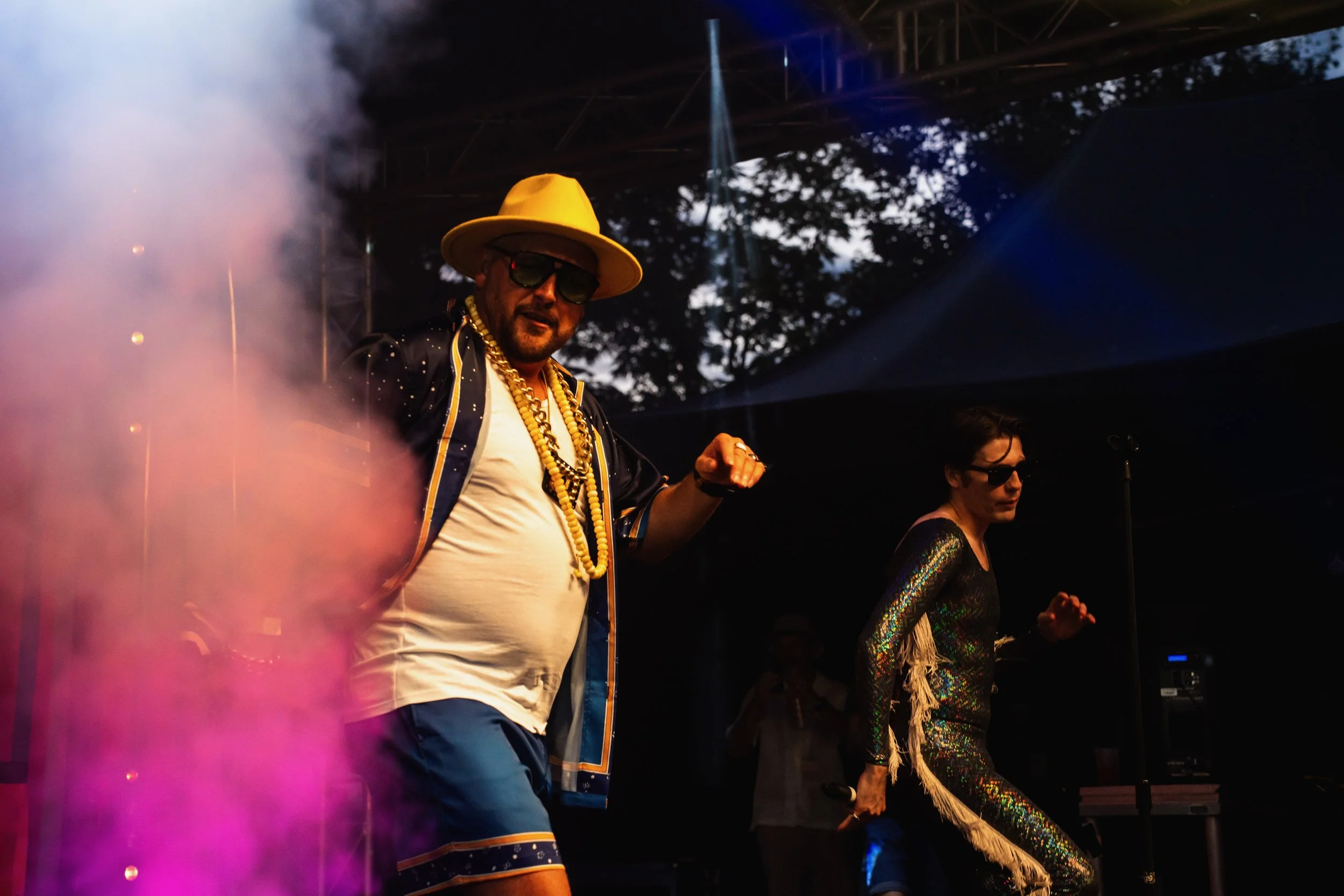 Two performers dancing on stage with colorful smoke, one in a wide-brimmed hat, sunglasses, gold chains, a patterned shirt, and shorts, the other in a sparkling dress with fringes and sunglasses, during a lively concert or event, outdoor setting with