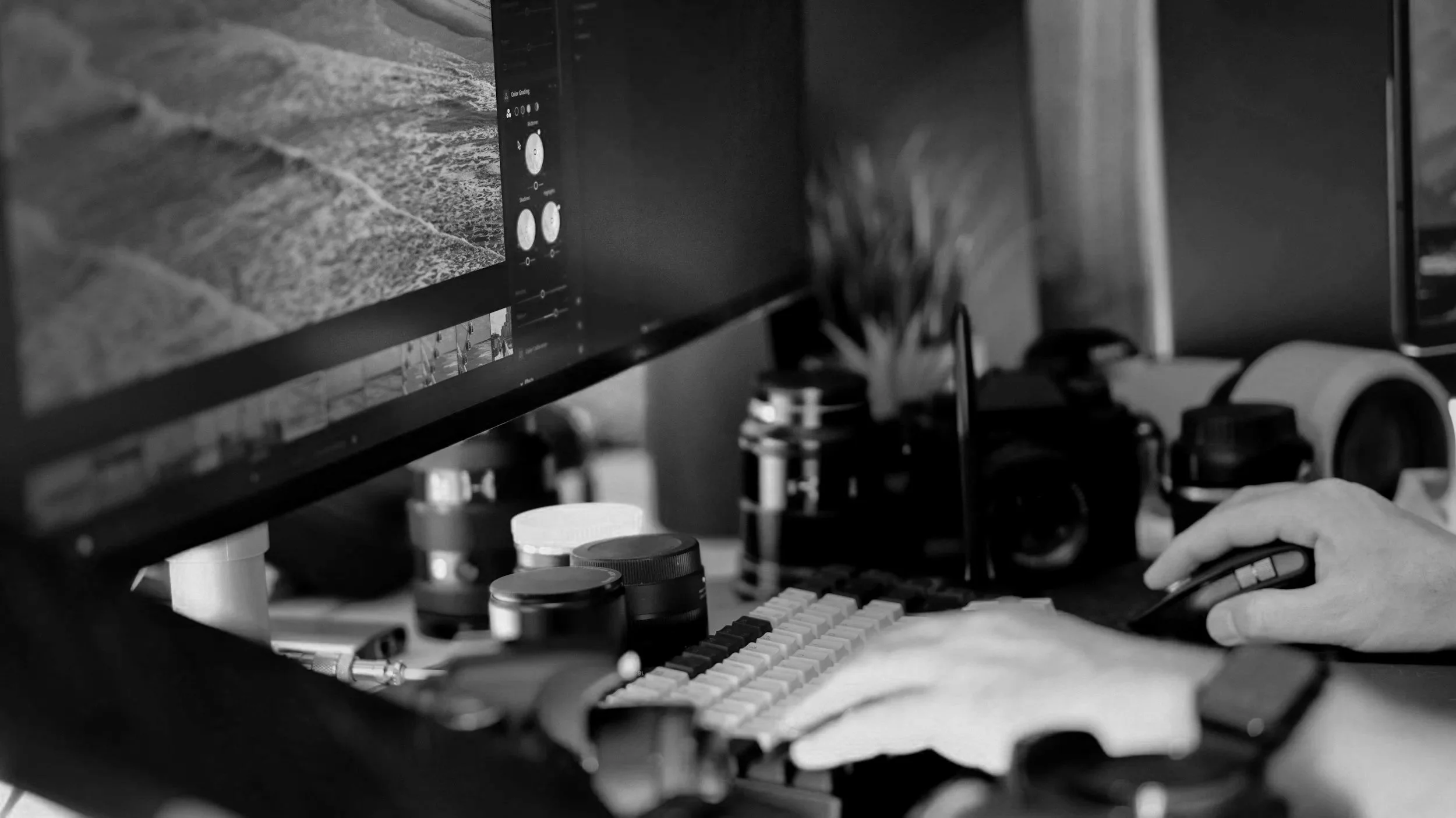 A person working on a computer with multiple monitors. The screen shows photo editing software. A keyboard, mouse, and various camera lenses and equipment are on the desk.