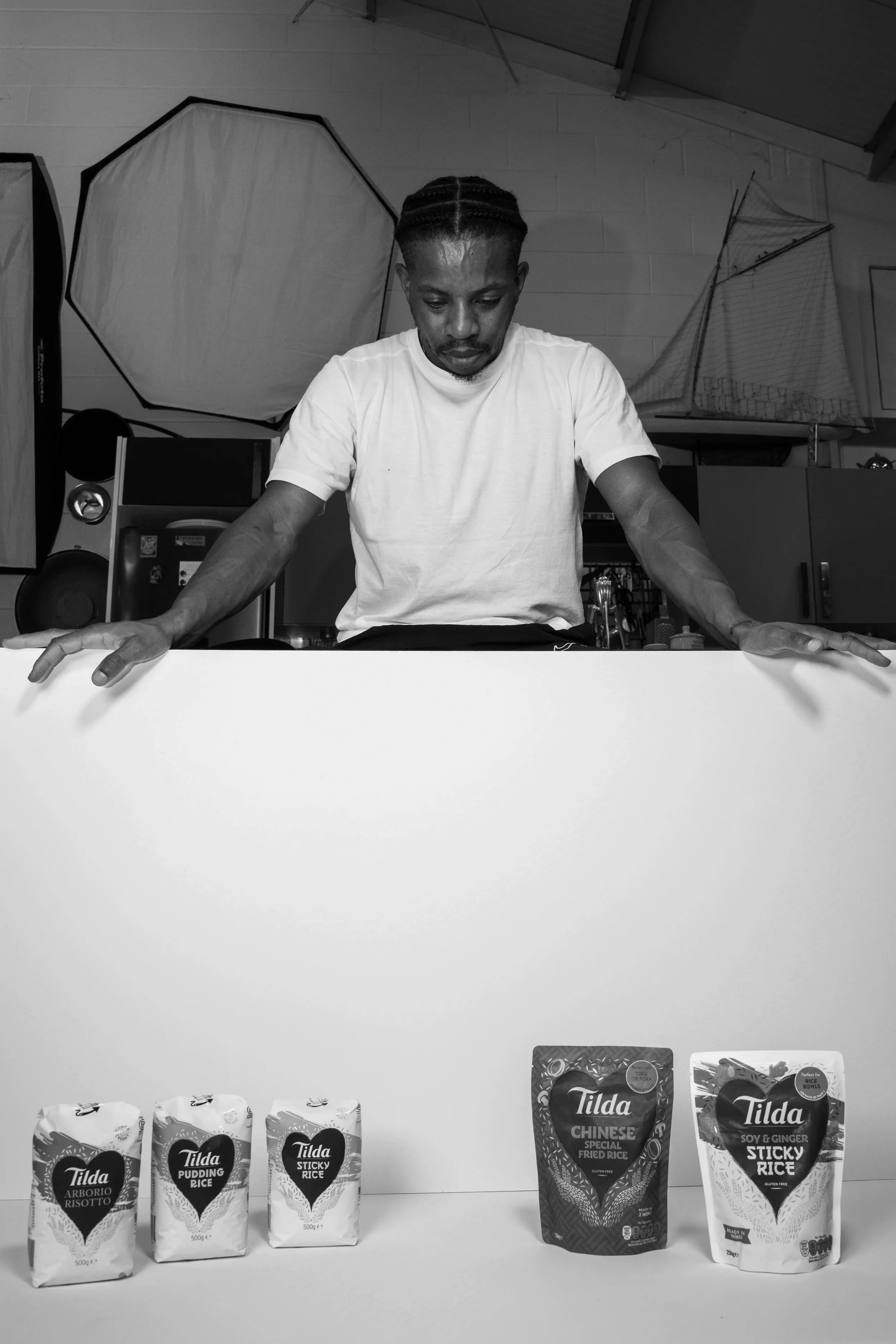 A man looking down at a row of rice packages on a table, with studio lighting equipment and a model ship in the background.